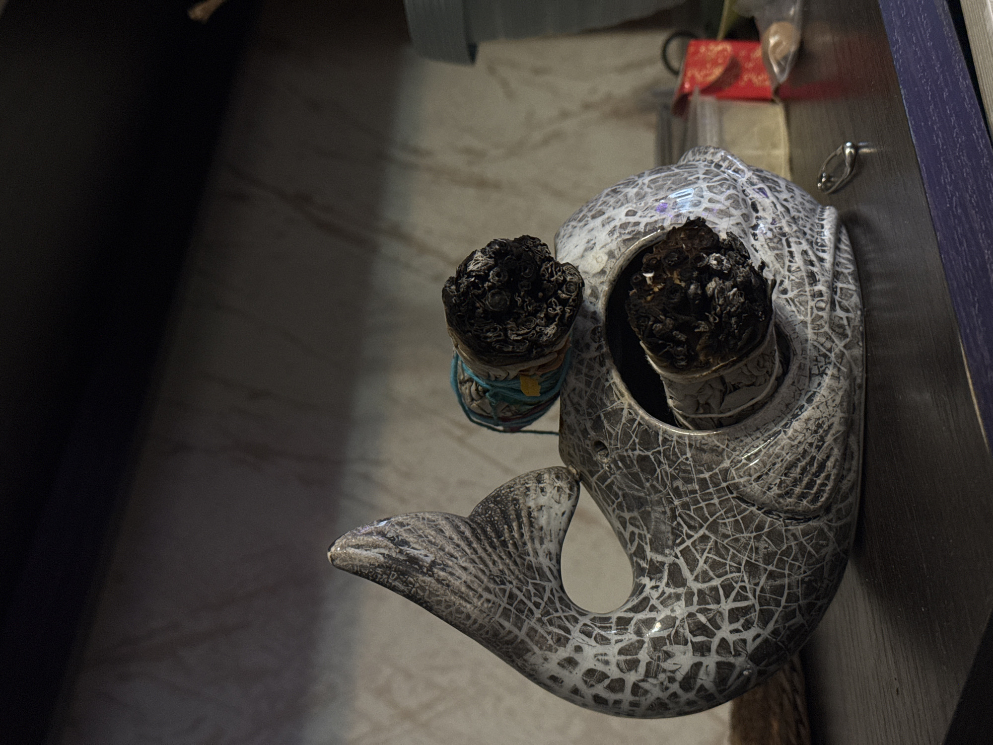 The image shows a decorative ceramic whale on a dark surface. The whale has a cracked, glazed finish in shades of gray and white. There are two bundles of charred material in the whale's "eyes." These appear to be spent smudge sticks or incense bundles. Other items can be seen in the background, including a red box and some other objects. The setting is not Kladovitsy, Russia.