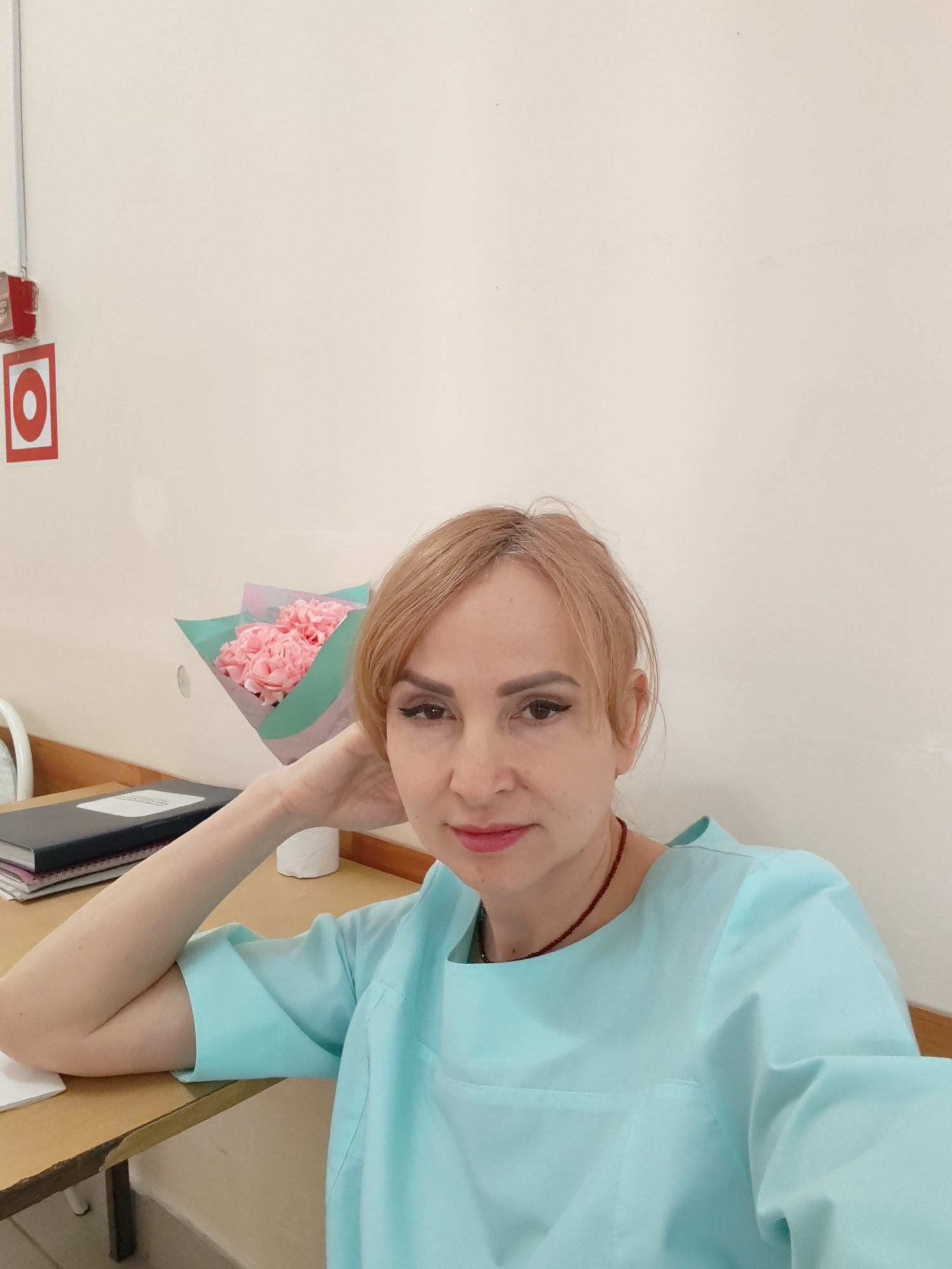 A woman is sitting at a desk indoors. She has blonde hair and is wearing a light blue scrub top and a red necklace. Her left arm is bent at the elbow and her hand is supporting her head. On the desk to her left are some notebooks and a vase with pink carnations. In the background, the wall is a light cream color and there is a red circle on a white square near the top left corner of the frame. The setting appears to be a medical facility, potentially in Chaikovsky, Russia.