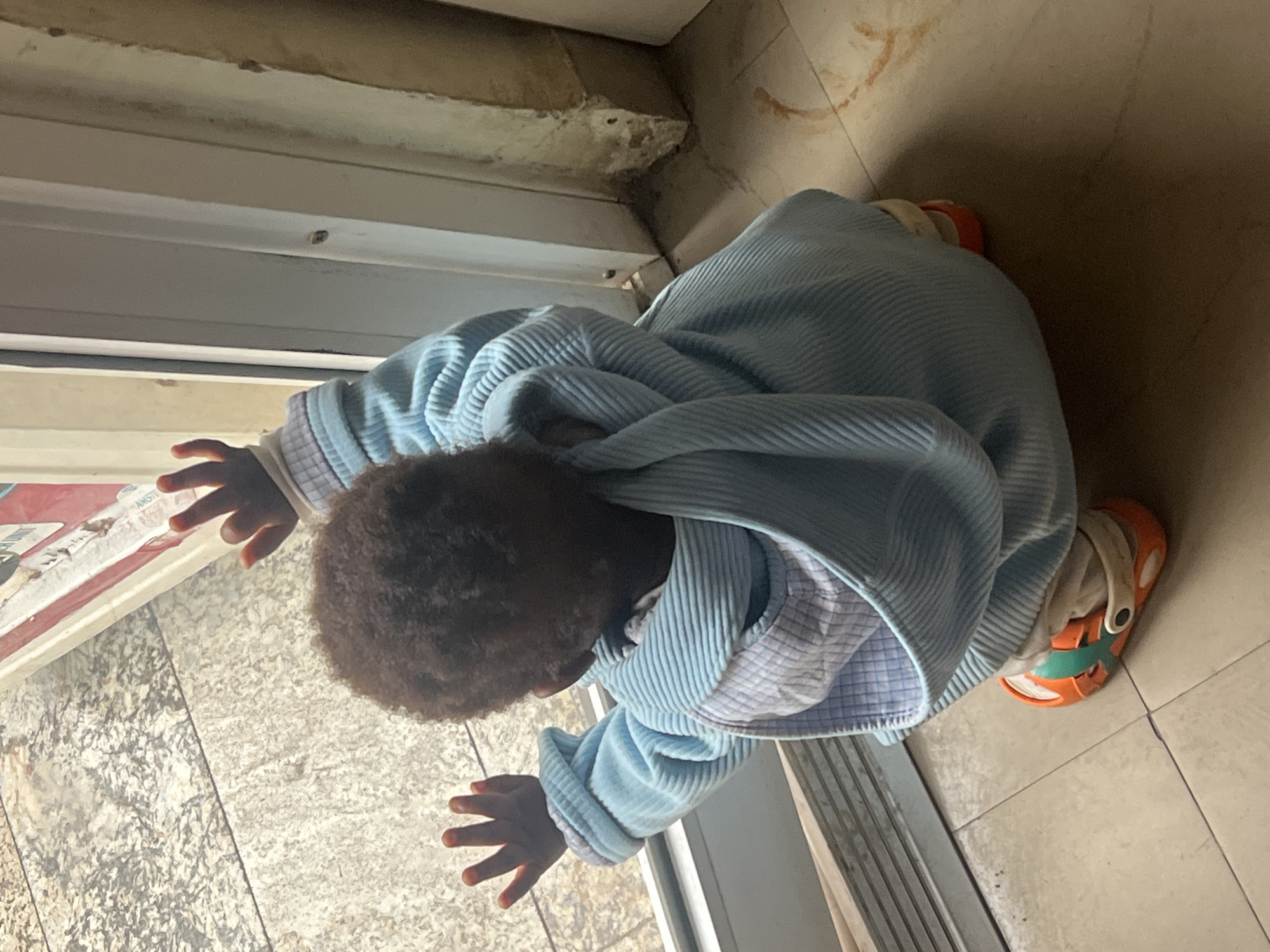 A child is observed indoors on tiled flooring in Gura Top, Nigeria. The child, with dark curly hair, wears a light blue corduroy jacket over a checkered shirt, and orange and white sandals. They are positioned at a window, with both hands pressed against the glass. The environment includes a white window frame and adjacent wall, with the tiles extending into the room. The scene is illuminated by daylight.