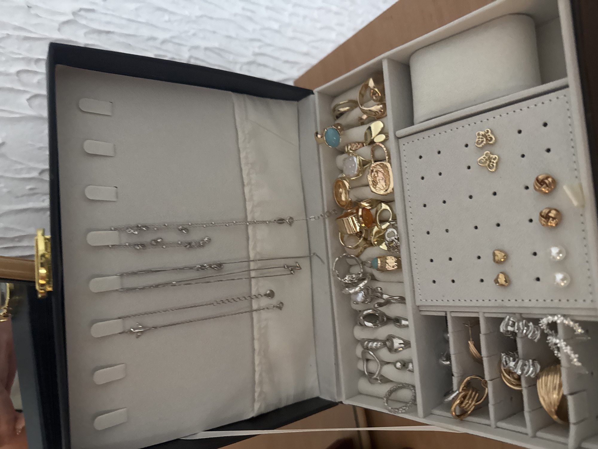 A partially opened jewelry box is displayed. The box has a black exterior and a white interior. The left compartment holds several silver-colored necklaces hanging from white tabs. The right compartment is divided into smaller sections containing a variety of jewelry, including gold and silver rings, earrings of various designs, and some items attached to a perforated display card. The background includes a textured white wall.