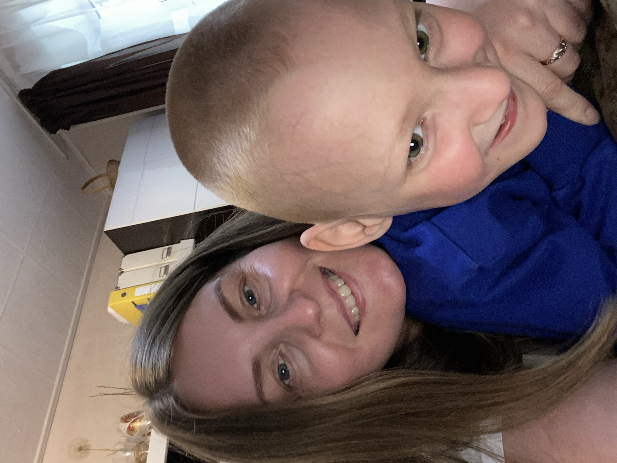 A selfie-style photograph shows a woman and a young child. The woman has light skin, blue eyes, and light brown hair. She is smiling. The child has a shaved head and green eyes. They are wearing a blue shirt or sweater. The location appears to be indoors. There are cabinets or a closet in the background. The location in the city of Noviy, Russia, is not discernible from the image.