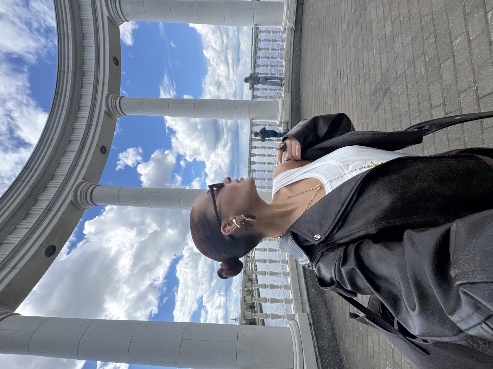 A woman stands on a paved walkway in Izhevsk, Russia. She is wearing a white tank top and a black leather jacket, a gold necklace, and dark sunglasses. Her dark hair is styled in a bun. A white colonnade with a rounded arch frames the scene, and beyond it, there is a view of a body of water with a railing. Two people can be seen standing at the railing in the distance. The sky is blue with white clouds.
