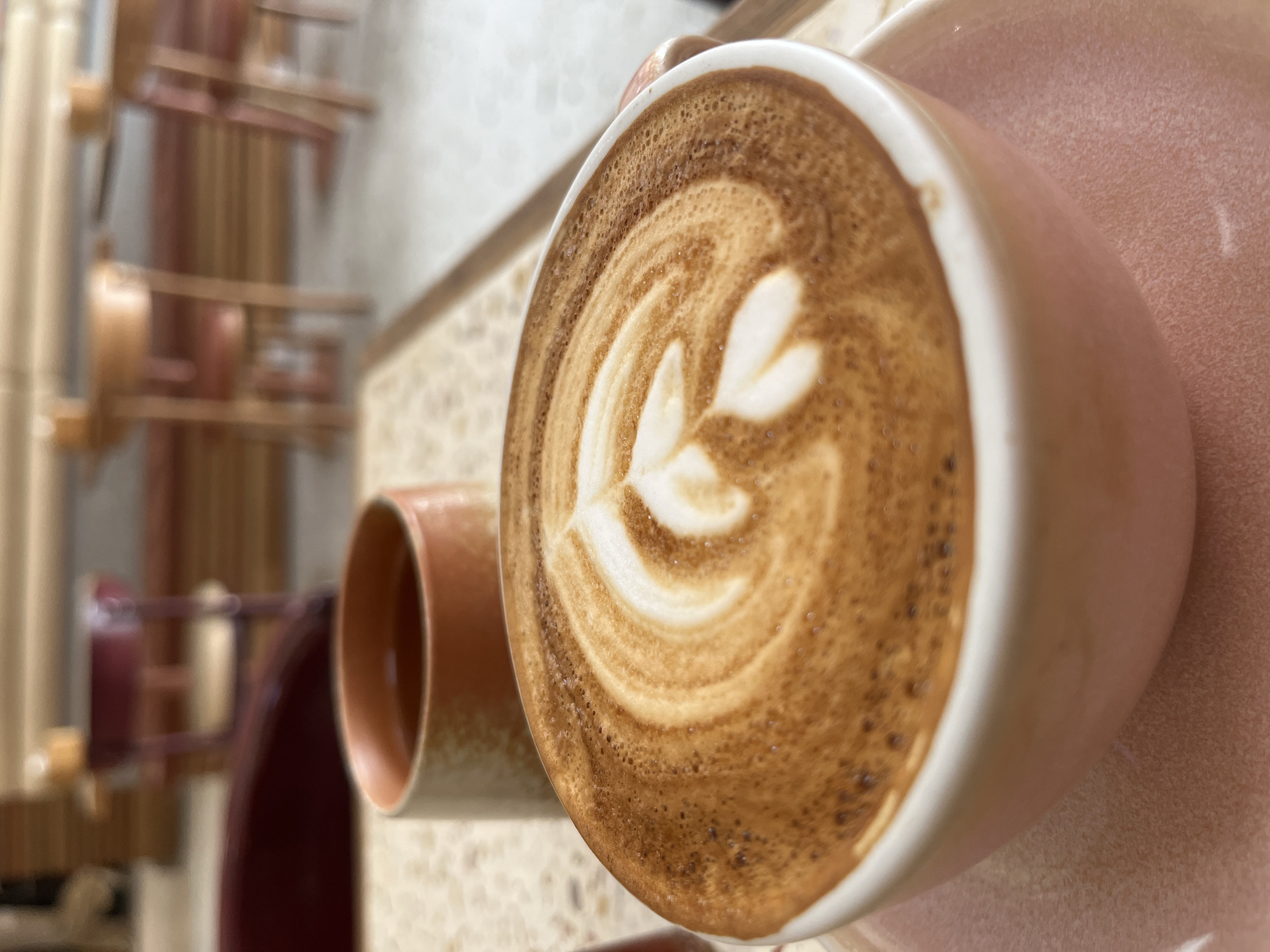 The image shows a cup of coffee with latte art on a saucer, likely inside a cafe or coffee shop. The coffee art features a leaf-like design. The cup and saucer are a pale pink color. In the background, out of focus, there appears to be a decorative wall arrangement with cylindrical shapes, suggesting a modern or artistic interior design. The location is possibly in Bac Nha Trang Ward, Vietnam.