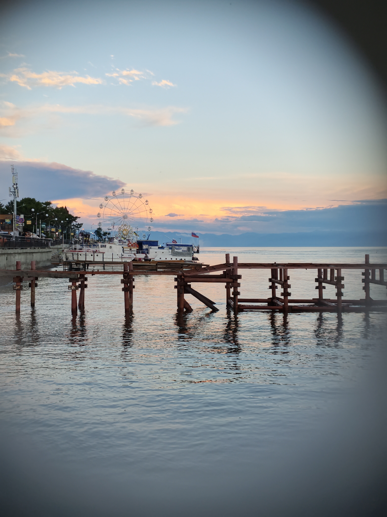 This image depicts a waterfront scene in Listvyanka, Russia, at dusk. In the foreground, an unfinished wooden pier with vertical supports extends into calm, rippling water, reflecting the sky and the structures beyond. The water is a cool blue-grey, with hints of orange and pink from the sunset. In the middle ground, several white boats are docked along a promenade. Behind the boats, a Ferris wheel with white spokes and cabins stands out against the colorful sky. The sky transitions from a pale blue at the top to shades of orange, pink, and purple near the horizon, with scattered clouds. A cellular tower is visible on the left side of the promenade, and trees line the background. The overall atmosphere is serene and slightly melancholic, characteristic of a quiet evening on a lake.