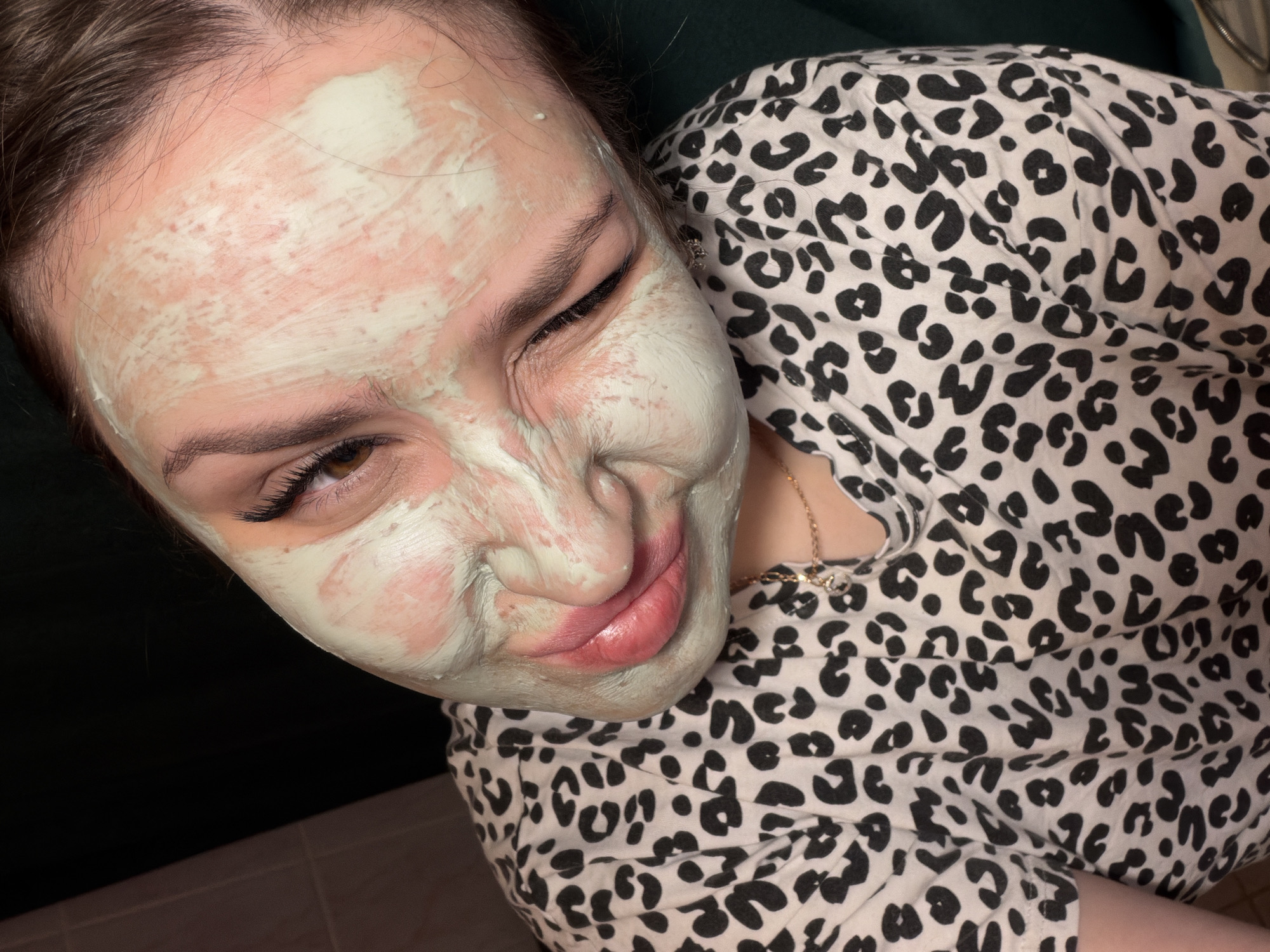 A young woman with fair skin is positioned at an angle, facing upward. Her face is partially covered with a light-colored facial mask, revealing her lips, left eye with dark eyelashes, and parts of her forehead and cheeks. She appears to be squinting. The woman is wearing a light-colored top with a black animal print pattern. The background includes a dark green surface and part of a tiled surface in the bottom right. The setting is likely indoors. There is no data to confirm this photo was taken in Chaikovsky, Russia.