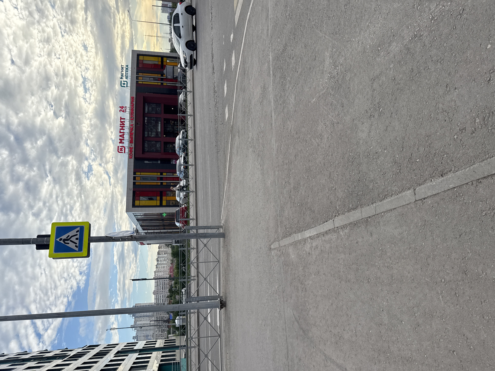 The image depicts a street scene in Perm, Russia, featuring a pedestrian crossing sign on a pole, a row of apartment buildings in the background, and a modern commercial building with storefronts. A white sedan is in motion on the right side of the road, and other vehicles are parked or moving in the background. The sky is cloudy, with patches of blue. The area appears to be a mix of residential and commercial urban development, with a visible pedestrian walkway and road infrastructure.