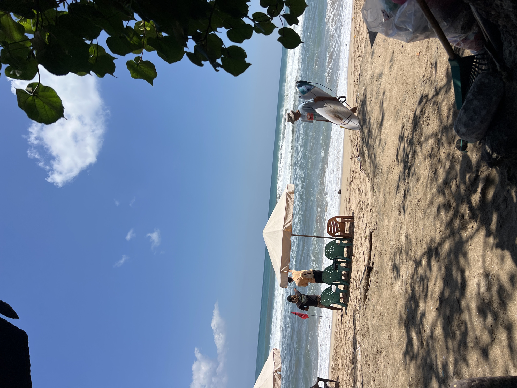 The image depicts a bright, sunny day at a beach in Kuta, Indonesia. In the foreground, the sand is textured with shadows from overhead foliage, and a trash bag, rake, and a large rock are visible. Further back, several beach chairs are set up under two beige umbrellas. Three people are present on the beach: two are standing near the water and an umbrella, one holding a red flag, and the other appears to be adjusting the umbrella. A third person, wearing a hat, walks along the shore carrying a surfboard. The ocean features moderate waves, and the sky is a clear blue with a few scattered white clouds.