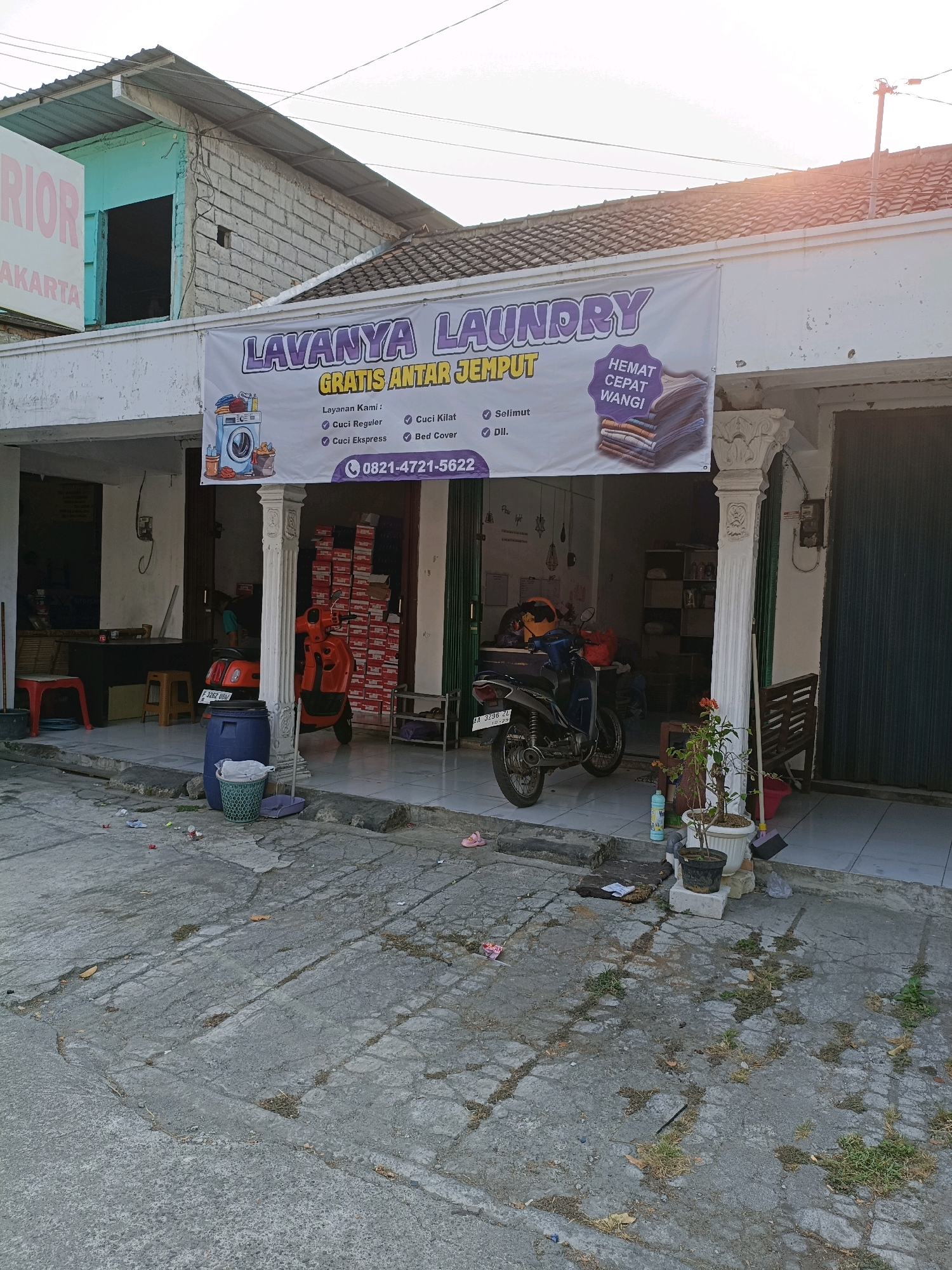 The image shows a street-level view of a laundry business located in Ngemplak, Indonesia. A large banner on the storefront reads "LAVANYA LAUNDRY" in purple text, with smaller text below indicating "GRATIS ANTAR JEMPUT" (free pick-up and delivery). The banner also lists services such as "Cuci Regular" (regular wash), "Cuci Kilat" (express wash), "Cuci Ekspres" (express wash), "Bed Cover", and "Selimut" (blanket), along with a phone number "0821-4721-5622". An illustration of a washing machine and folded clothes is on the left side of the banner, and a stack of folded clothes with the text "HEMAT CEPAT WANGI" (economical, fast, fragrant) is on the right.
In front of the laundry, there is a blue trash bin and a green trash basket. Two motorcycles are parked in front of the business, one orange and one black. The orange motorcycle is partially obscured by a white pillar. To the left of the entrance, there are two red stools and some stacks of what appear to be boxes. Inside the laundry, shelves are visible. The building appears to be a single-story structure with a tiled roof. The ground in front of the business is paved with concrete that shows signs of wear and cracking, with some patches of grass growing through. There are also various items scattered on the ground, including small pieces of trash, a flip-flop, and a bottle of laundry detergent. The overall environment suggests a typical urban street scene in Indonesia.