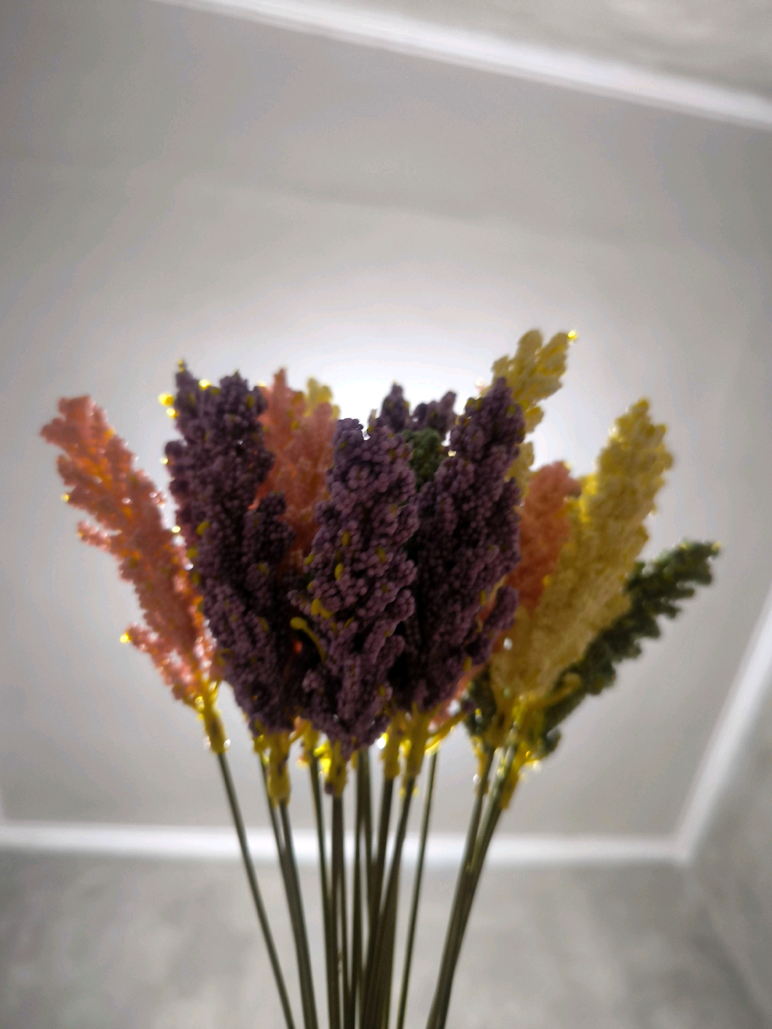 A close-up, low-angle shot shows a bouquet of dried, colorful flowers against a white ceiling. The flowers are arranged in a fan shape, with stalks extending downwards out of frame. The blooms themselves are dense and textured, resembling small feathery plumes. They are colored in shades of pink, purple, yellow, and green, with some specks of glitter. The lighting appears to be from above, creating a soft glow and some highlights on the flowers. The background consists of a textured, off-white surface that suggests a wall or another surface, with a subtle shadow or gradient. The image has a shallow depth of field, with the flowers in the foreground being the sharpest elements. The scene is indoors, likely in a domestic setting, and does not contain any discernible elements that would indicate a specific location in Tabanan, Indonesia.
