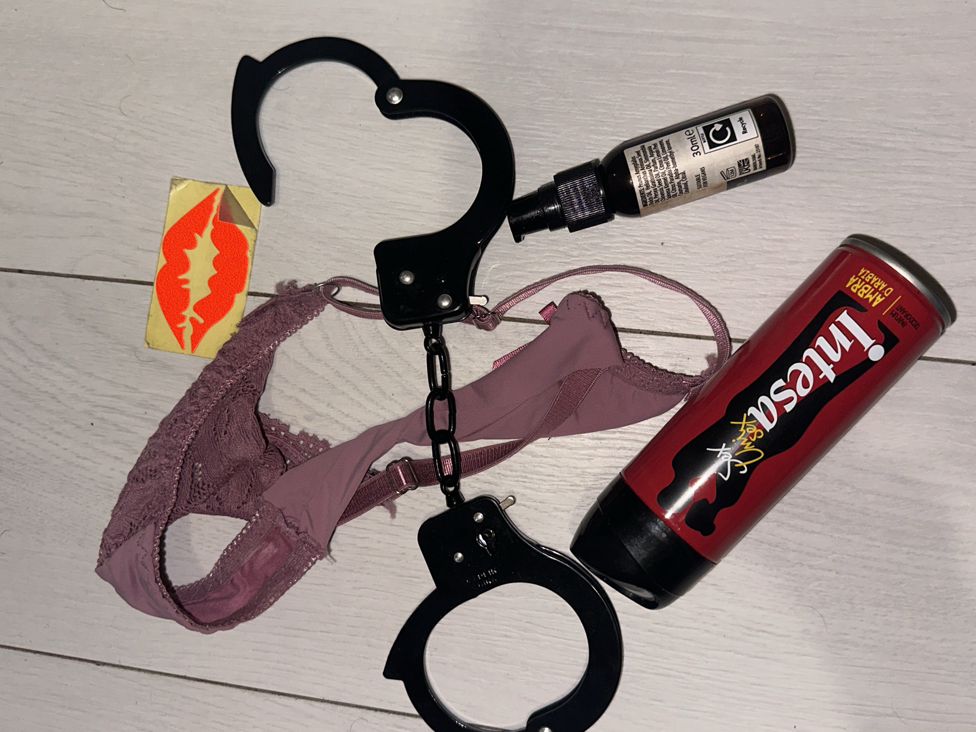 The image displays an overhead view of various objects arranged on a light-colored wooden surface, possibly flooring, exhibiting visible grain.
Central to the composition is a pair of dark mauve or purple lace-trimmed underwear, appearing to be a thong or G-string style, with a visible light-colored stain in the crotch area. A black metal pair of handcuffs, connected by a short chain, is positioned around the underwear. One cuff is open, while the other is closed. The strap of the underwear is threaded through the chain linking the handcuffs.
Above and slightly to the right of the underwear is a small, rectangular, light yellow card or paper featuring a prominent, glossy red graphic of stylized lips with a silver-colored highlight.
In the lower-left corner of the frame, a red aerosol can with a black cap is present. It features a black silhouette design of two figures and the brand name "Intesa" in white and yellow lettering. Text below indicates "AMBRA D'ARABIA" and "PARFUM DEODORANT."
In the lower-right quadrant, a small, dark brown glass bottle with a black pump dispenser is visible. Its label lists ingredients including various oils (Prunus Amygdalus Dulcis Oil, Helianthus Annuus Seed Oil, Persea Gratissima Oil, Simmondsia Chinensis Seed Oil, Argania Spinosa Kernel Oil, Citrus Limon Peel Oil, Citrus Paradisi Peel Oil), Parfum, and other compounds. The label also specifies "30mle," "SUITABLE FOR VEGANS," "UK Made," "Recycle," and "Batch No: 23187."