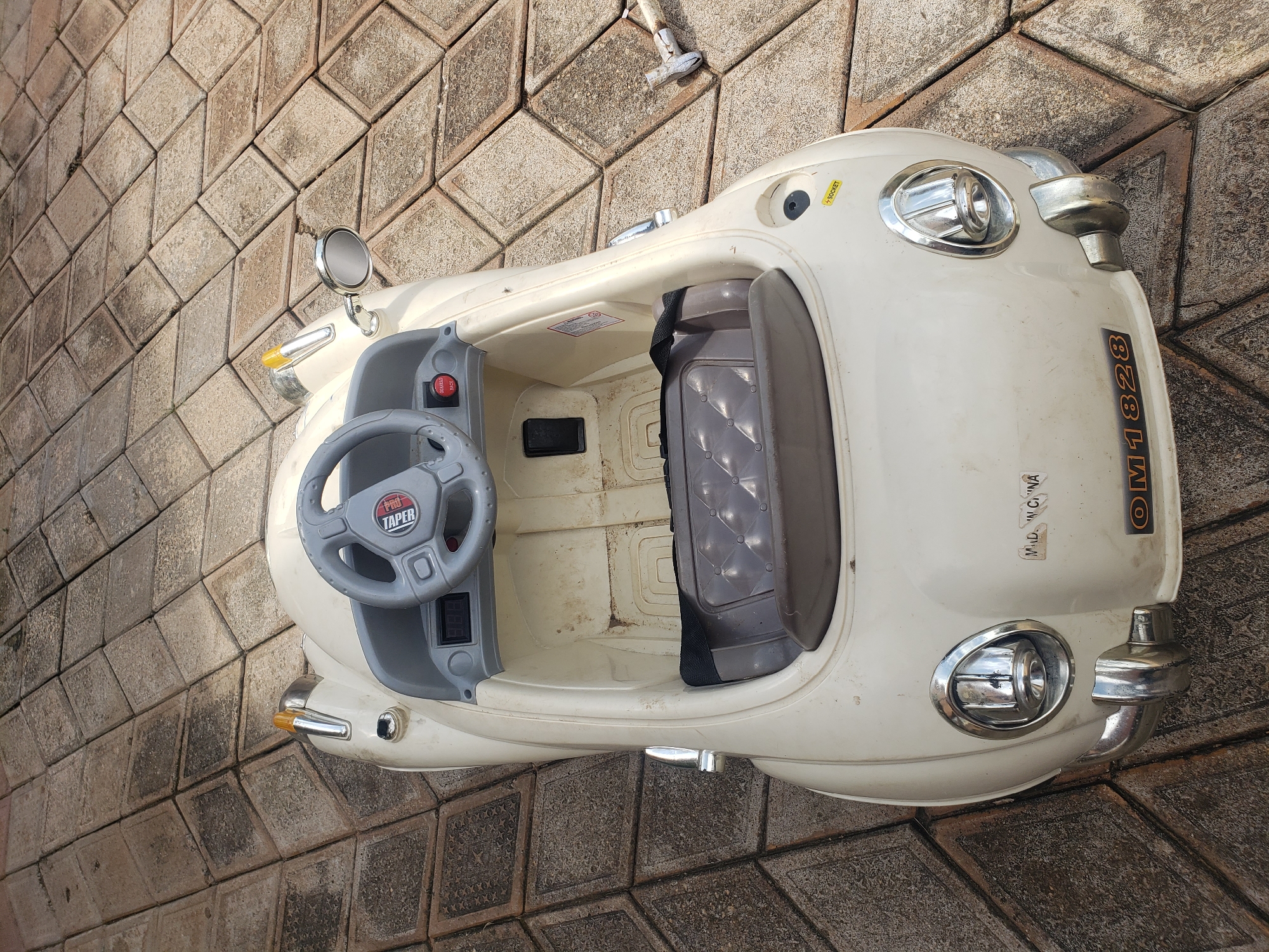 An overhead, high-angle shot captures a white toy electric car parked on a textured, outdoor patio paved with light-brown, square tiles. The car, designed to resemble a vintage convertible, is slightly dusty and appears to be weathered. The interior of the car features a gray steering wheel with a red and white logo, a digital display, and a black seat with a quilted pattern. A small red button is visible on the dashboard. Stickers adorn the car's exterior, including "MADE IN CHINA" and "OM1828" on the front. A chrome-plated side mirror extends from the passenger side of the car. The patio is uneven, with some tiles showing wear and discoloration. The car is positioned in the center of the frame, angled slightly towards the bottom right corner. The overall scene suggests the car is in a residential area, possibly a backyard or driveway in Sabon Tasha, Nigeria.