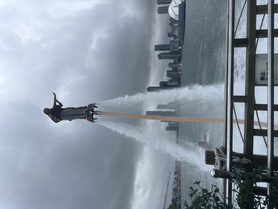 This image depicts a water performance in Qingdao, Shinan District, China, under a heavily overcast sky. Two individuals are engaged in a tandem flyboard maneuver, elevated high above the dark, choppy water by twin powerful jets of water emanating from the flyboard's foot bindings. The primary operator, wearing light-colored trousers and a dark top, stands on the board while supporting a secondary individual, who is positioned in front and extends their left arm horizontally. A reddish-brown pole or hose extends vertically from the flyboard assembly. In the mid-ground, a red personal watercraft, likely powering the flyboard, is partially visible. The background showcases the modern skyline of Qingdao, featuring numerous high-rise buildings and a distinctive white dome structure on the right. To the left, a pier or waterfront area is visible, adorned with a long line of diverse flags. The foreground includes a wooden railing with metal bars and a sign bearing Asian characters, alongside some green foliage.