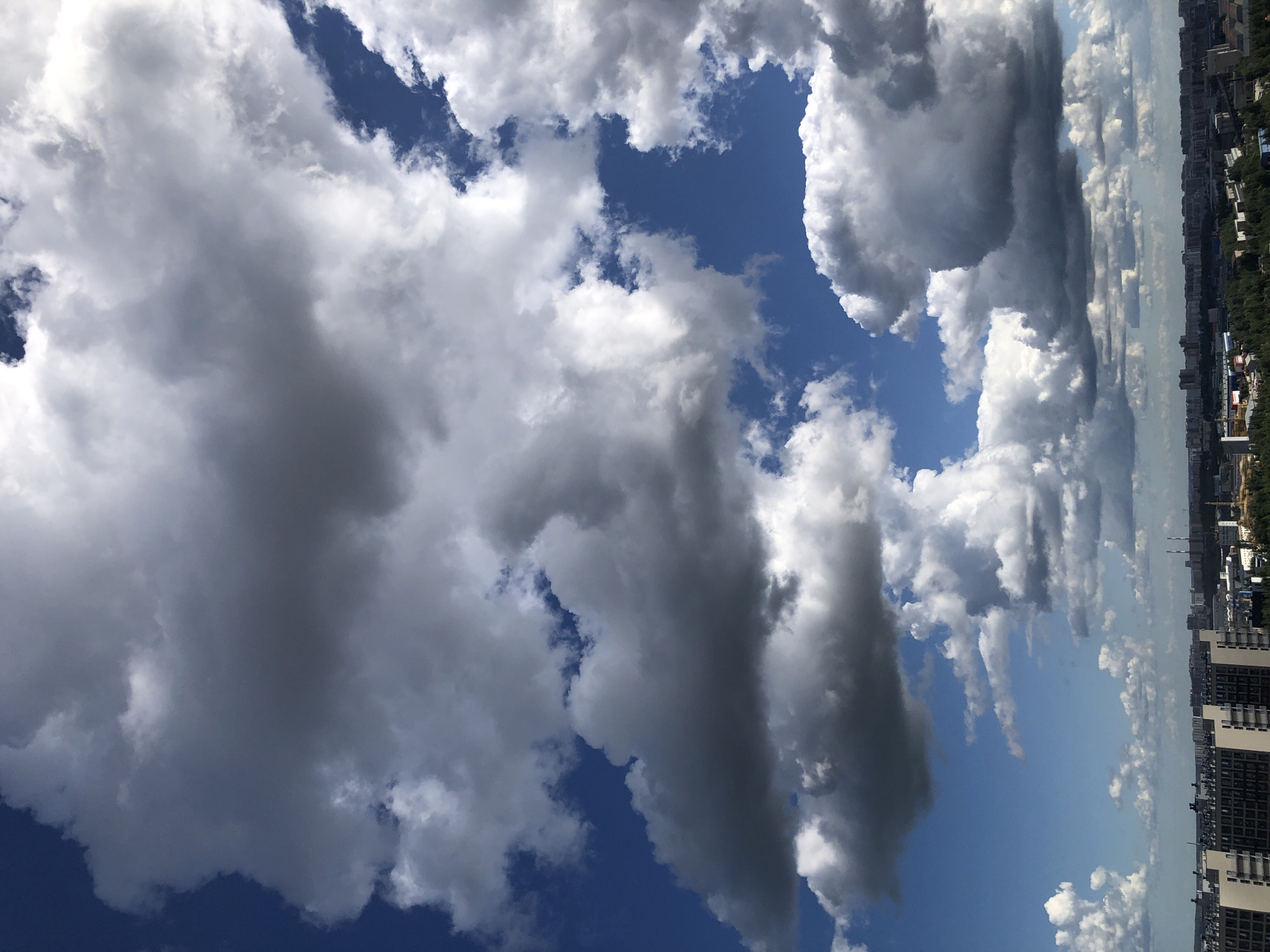 The image captures a panoramic view of a city skyline under a dynamic sky filled with dramatic clouds. The lower portion of the image displays a dense urban landscape with buildings of varying heights and architectural styles, interspersed with patches of green trees. The city extends into the horizon, with some structures appearing taller and more industrial, including what appear to be smokestacks in the distance. Above the city, the sky is a vibrant blue, punctuated by a variety of cloud formations. Large, voluminous cumulus clouds dominate the upper and central parts of the frame, some casting dark shadows, suggesting they are dense and possibly holding moisture. Other, smaller clouds are scattered across the sky. The lighting suggests it is daytime, with the sun illuminating the white parts of the clouds and casting shadows on the darker, gray portions. The overall impression is of a slightly overcast but bright day over a sprawling city.