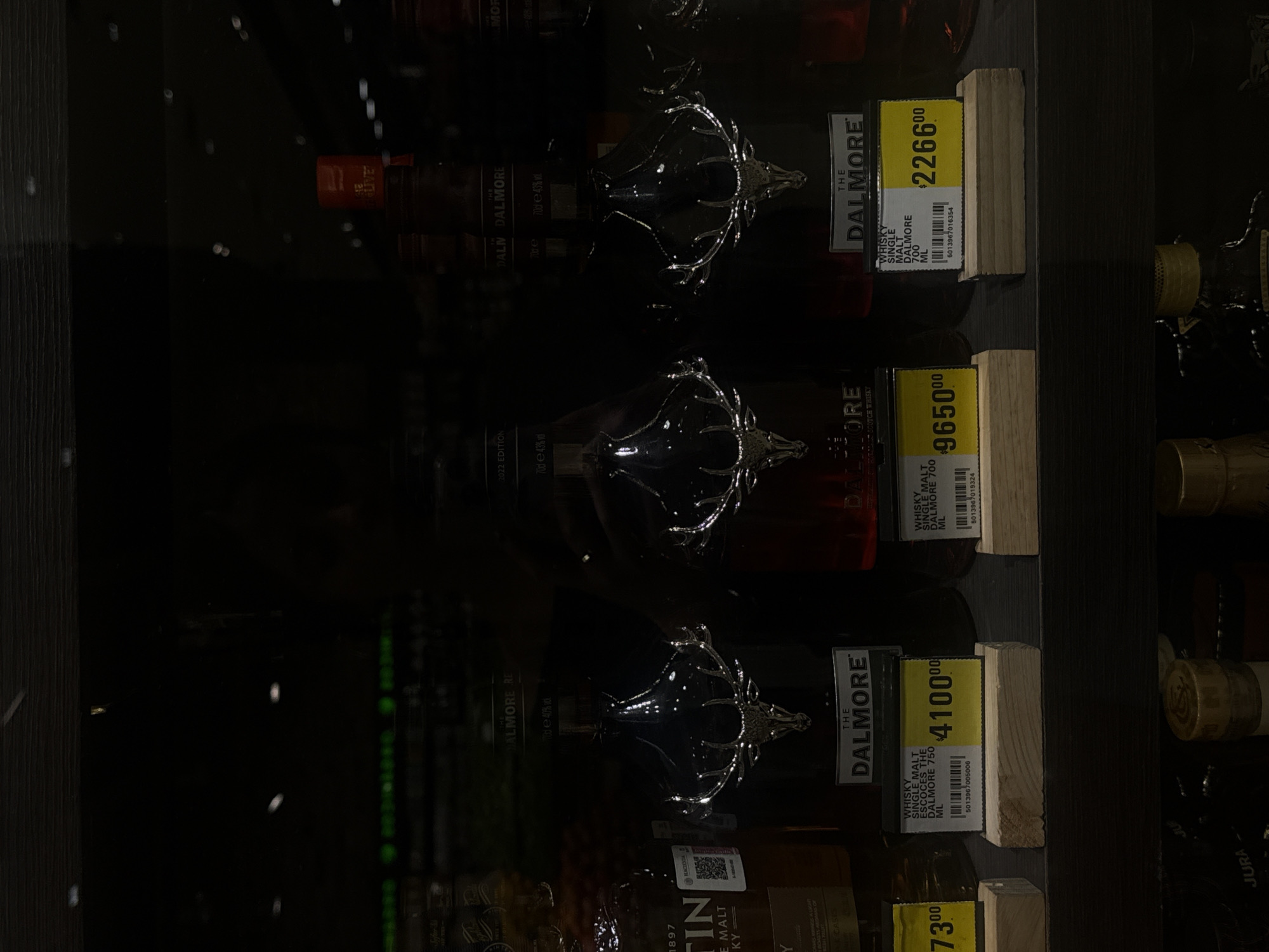 The image depicts a shelf in what appears to be a retail establishment, likely a liquor store, possibly in the city of Unknown, Unknown. Displayed on the shelf are several bottles of "The Dalmore" single malt Scotch whisky. Price tags are prominently displayed next to each bottle, indicating the price of the products: $2266.00, $9650.00, and $4100.00. The bottles feature distinctive deer head emblems. The lighting in the store is dim, creating a reflective surface on the bottles. Other bottles are visible on the right side of the frame.