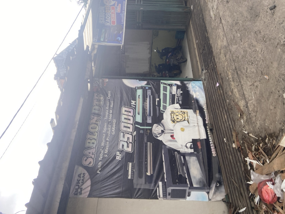 The image shows a storefront with a large banner advertising "Sablon DTF" printing services in Baleendah, Indonesia. The banner features a large price of "RP 25.000" for printing, and showcases various printing equipment, including three large printers. The banner also advertises "Putih Tebal - Warna Tajam - Tanpa Antri - Cepat - 5 Mesin Produksi - 1000 M/Hari - Garansi." The storefront is partially obscured by the banner and there is a sign on the right side of the image advertising "Sablon Kaos DTF" and prices. Two motorcycles are parked on the left side of the storefront, and a doorway leads into the shop. There is a pile of debris in the foreground of the image, directly in front of the storefront. The location of the storefront is identified as Baleendah, Indonesia with the coordinates -7.005 latitude and 107.631 longitude. The sky is partly cloudy and the ground is a mixture of concrete and dirt. There are no people visible in the image.