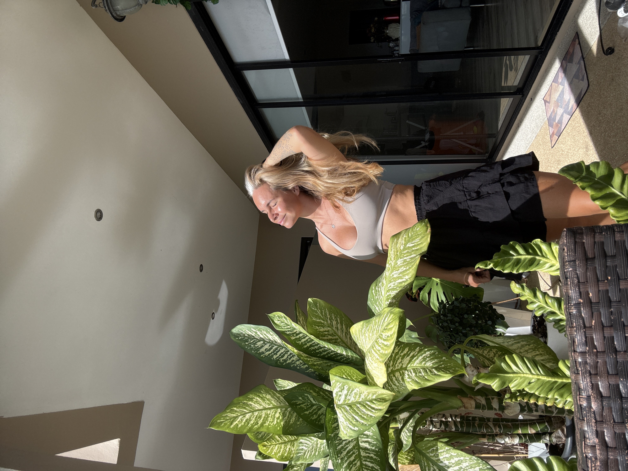 A woman with blonde hair stands in front of large green plants. She wears a light-colored sports bra and a black ruffled skirt. The woman has her eyes closed and her hand on her head, basking in sunlight. The background shows a building with large glass doors and a portion of an outdoor patio with a decorative mat on the ground. The image appears to be taken outdoors, possibly on a balcony or patio in Baan Chaweng, Thailand, as indicated by the tropical foliage and architectural style.