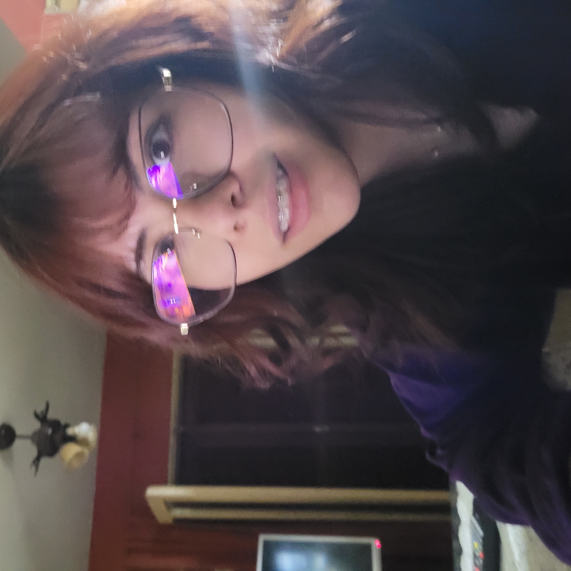 A young woman with light brown hair and wearing glasses is the primary subject of this selfie-style image. She has braces on her teeth and is smiling.
Behind her, in the background, is a room with a red wall, a television, and a ceiling fan. These elements suggest the interior of a home. No specific location in San Fernando del Valle de Catamarca can be determined from the image.