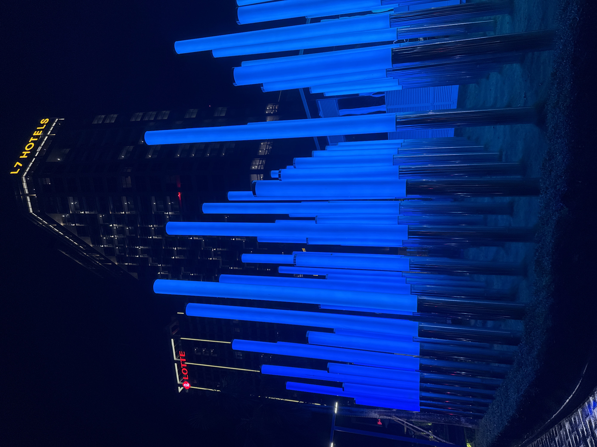The image shows a night scene in Hanoi, Vietnam, featuring an art installation of blue illuminated cylindrical lights in the foreground, arranged in rows and varying in height. Behind the lights, two tall buildings are visible. The building on the left has a red illuminated "LOTTE" logo and vertical yellow lights. The building on the right is taller and displays a yellow illuminated "L7 HOTELS" sign at its top. The overall ambiance is modern and urban, with the lights creating a striking visual contrast against the dark night sky and building architecture.