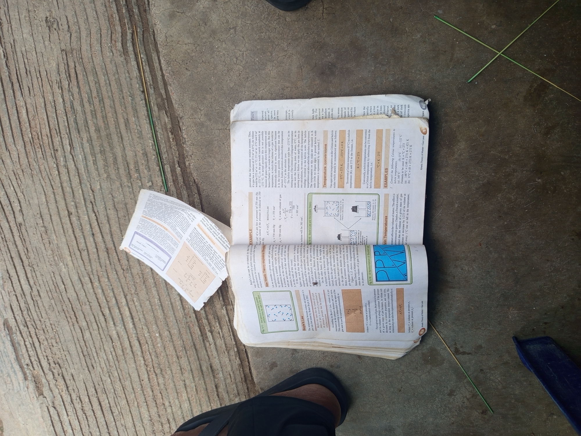 The image depicts an open textbook lying on a textured, grey concrete surface, possibly a pavement or ground. The book appears to be a science textbook, with diagrams and text related to physics, specifically kinetic theory and gas laws. Some loose papers, also with printed text, are placed on top of the textbook. The scene is lit by natural light, casting faint shadows. Two green straws are present on the concrete surface, one lying straight and another bent at an angle, crossing the first. A sliver of a dark object, possibly a shoe or sandal, is visible on the left side of the frame, suggesting the presence of a person. The overall environment is outdoors, with the concrete surface suggesting an urban or semi-urban setting. No specific landmarks or indications of Jalingo, Nigeria are visible within the image itself; the setting is generalized to an outdoor concrete area.