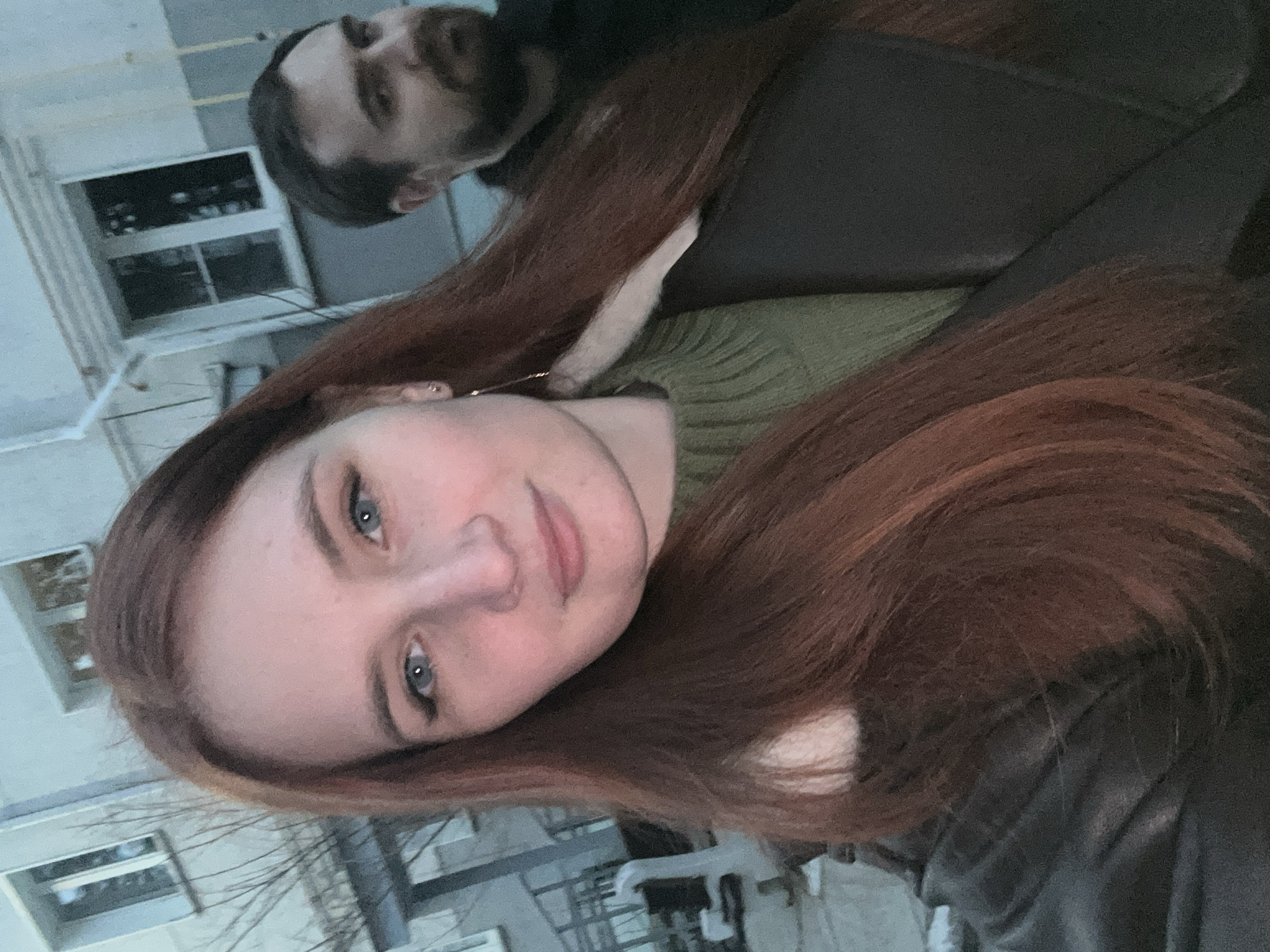 Two people are in the image. The primary focus is a woman with red hair, fair skin, and blue eyes. She is wearing a green sweater and a black jacket. Her face is angled towards the camera. Behind her, out of focus, is a man with a beard. A building with white frames is visible in the background. The exact location in the city of Chaikovsky, Russia is not definitively identifiable based on the provided image.