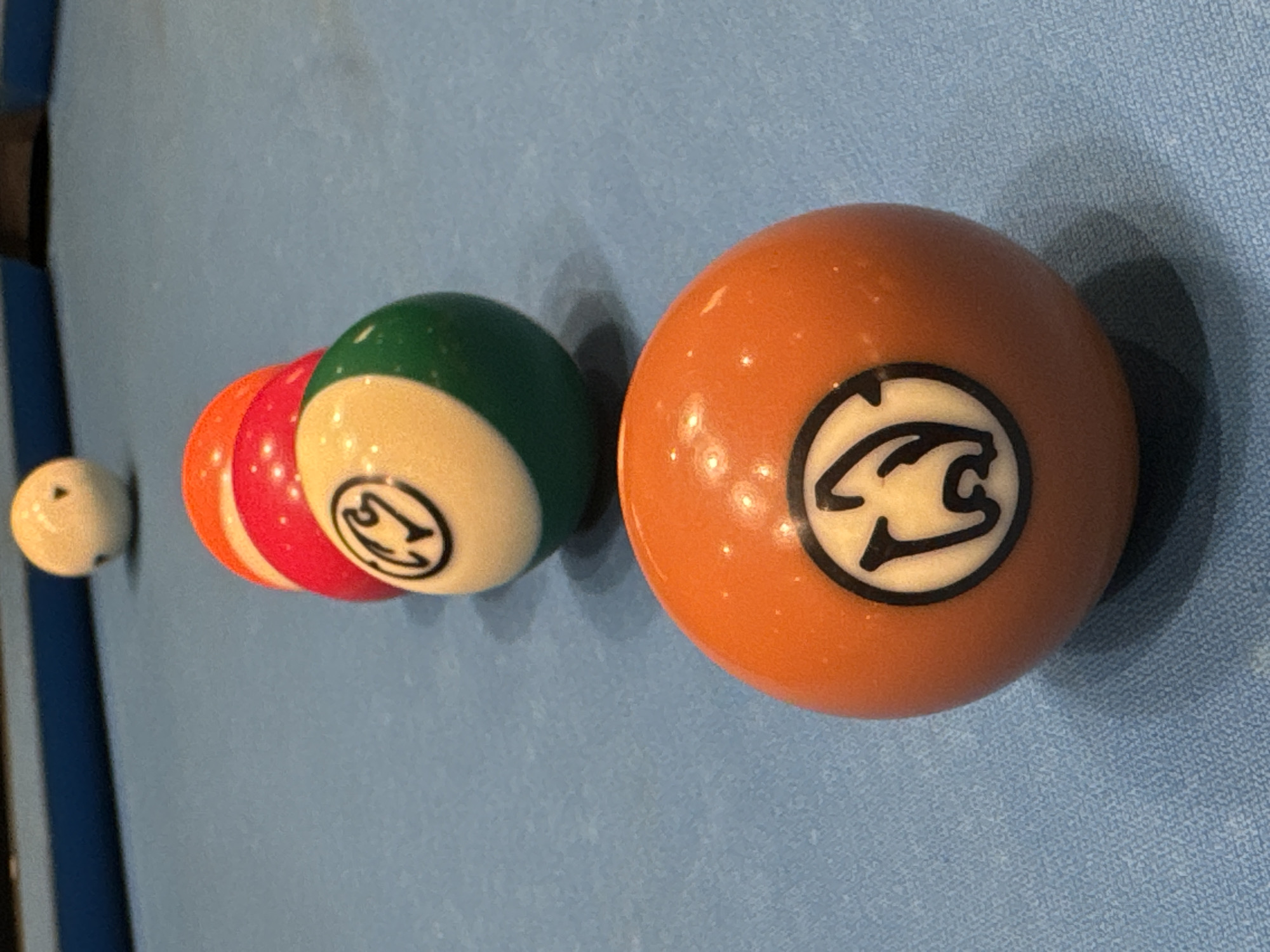 The image shows a close-up of a billiard table. Five billiard balls are visible in a row. The first ball is white. Then there is an orange ball followed by a red ball. Next, there is a green and cream colored ball, and finally an orange ball with a graphic logo on it. The table is covered in light blue cloth. There is no evidence to indicate a location in the city of Bellville, South Africa, or to suggest the presence or actions of any people.