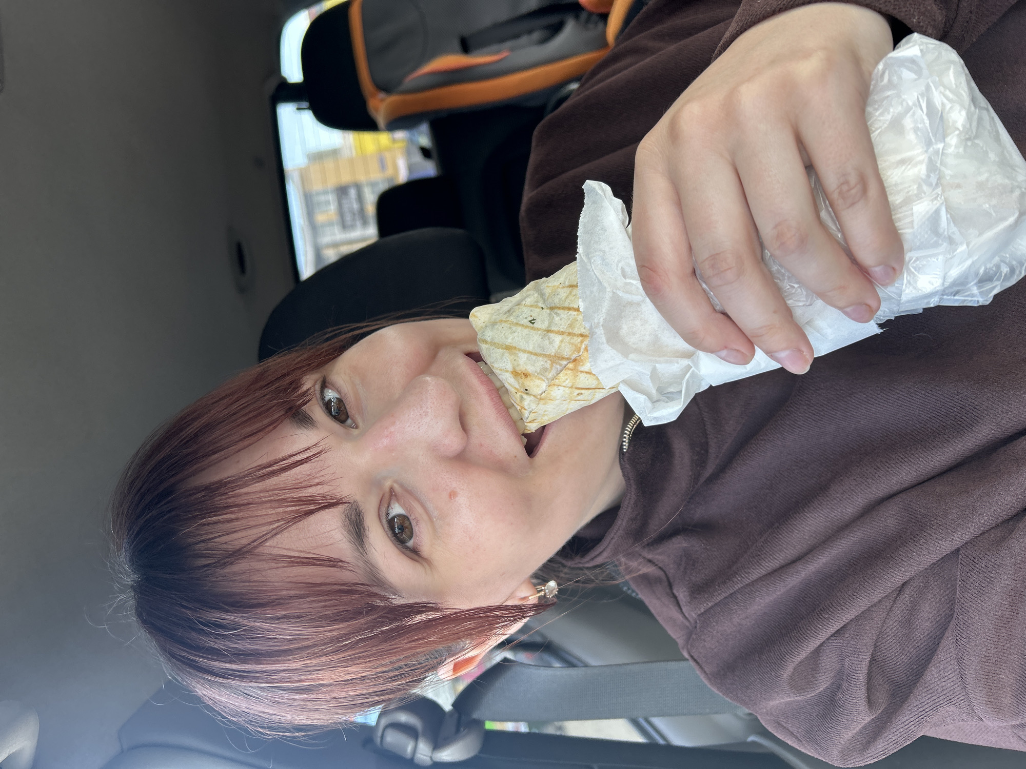 This image captures a close-up, rotated view of a person (likely female) inside what appears to be a car. The individual, with reddish-brown hair styled with bangs, is wearing a brown, ribbed long-sleeved top. They are actively engaged in eating a food item, which resembles a grilled wrap or panini, partially unwrapped from white paper and clear plastic packaging. The person's mouth is open, mid-bite, and their eyes are looking directly towards the camera, suggesting a candid or selfie-style shot.
The setting is inside a vehicle, with parts of the car's interior visible, including a black and orange car seat or accessory, and seatbelts. Through the car window in the background, vague shapes of distant buildings and an outdoor scene are discernible, indicating it is daytime. The natural light highlights the details of the person's face and the food. The overall mood is casual and spontaneous, depicting an on-the-go meal. There is no visible text on any of the objects or surfaces within the image.