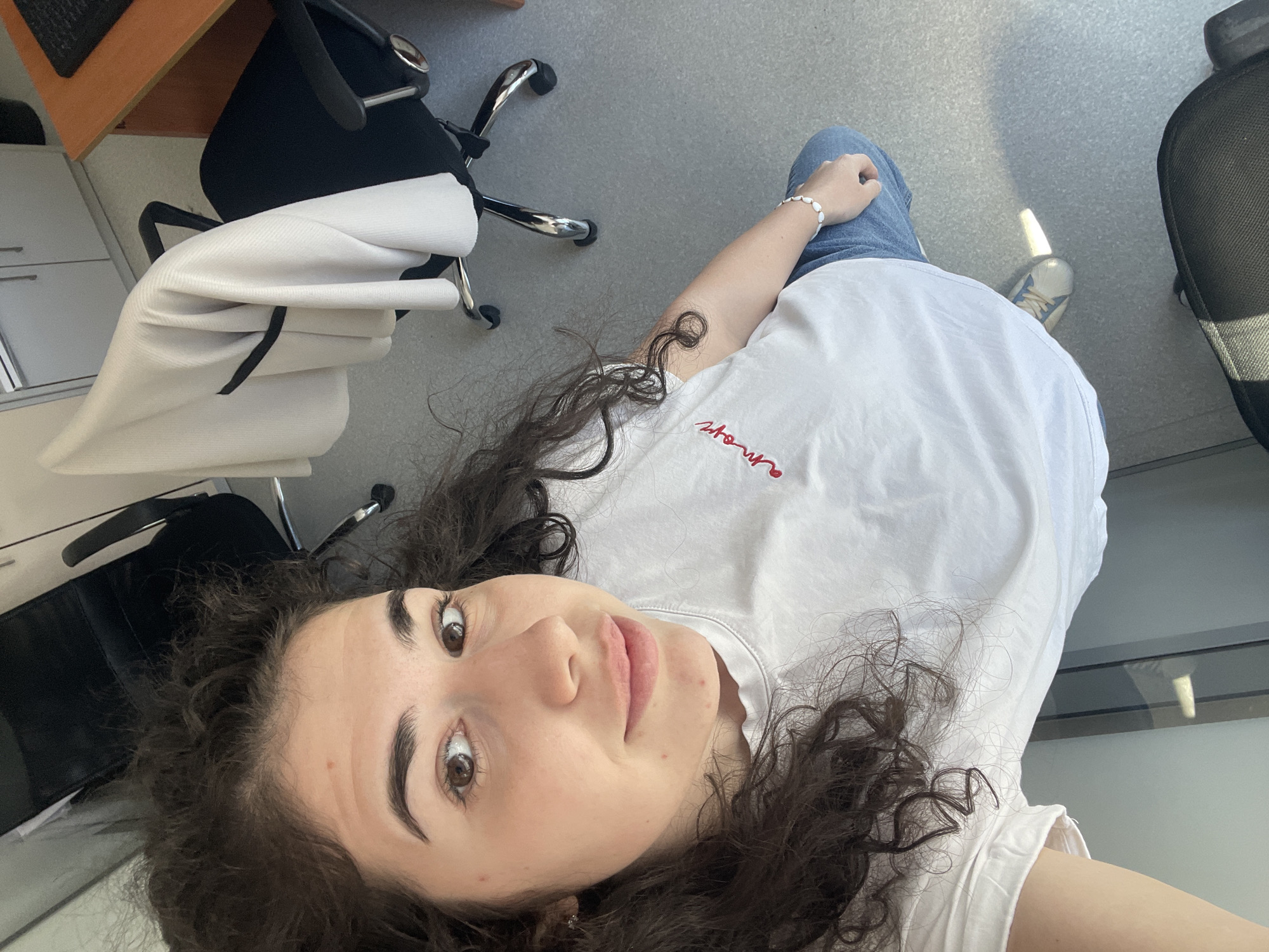 A high-angle, first-person perspective selfie captures a young woman looking up into the camera. She has dark, curly hair, light brown eyes, and defined eyebrows, with a calm and gentle expression. She is wearing a white t-shirt with "amour" embroidered in red cursive text on the chest, blue jeans, and white sneakers with light blue accents. A white beaded bracelet is visible on her left wrist.
The setting appears to be an indoor office or workspace. The background features a light grey floor, a black office chair with chrome legs and wheels, and a white cabinet or drawer unit. A light-colored wooden desk is partially visible in the upper left. Strong natural light, likely sunlight, enters from the right, illuminating the scene and casting some shadows on the floor, suggesting it is daytime. The overall scene is bright and casual, depicting a momentary pause in an office environment, possibly in Sofia, Bulgaria.