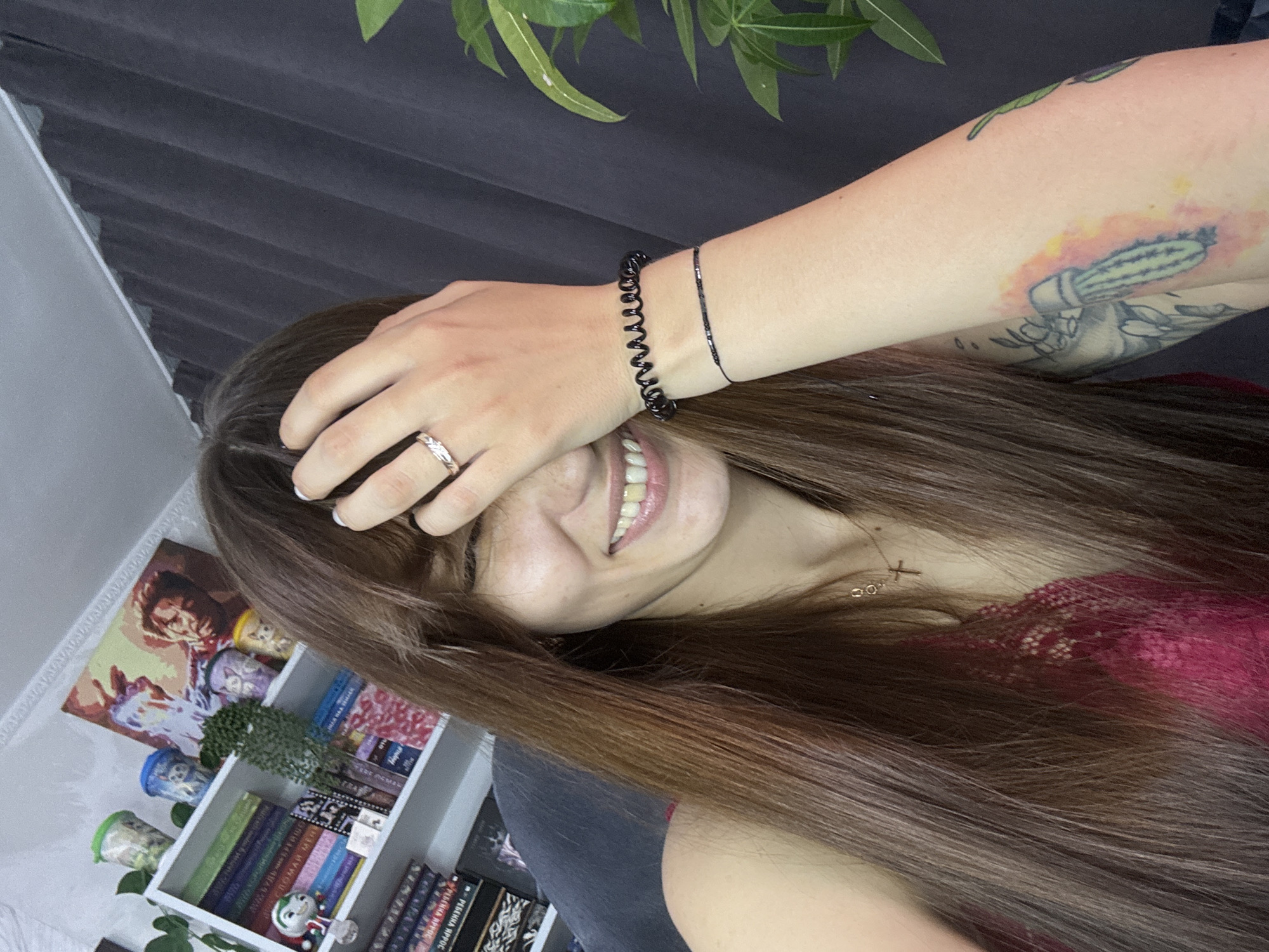 A young woman with long brown hair is smiling and covering part of her face with her right hand. She is wearing a red lace top and a necklace with a cross pendant. A ring is visible on her ring finger. She has a tattoo on her right arm that appears to be a cactus. Behind her, a bookshelf is filled with books and decorative items. The scene suggests an indoor setting, possibly a home in Novokuznetsk, Russia, given the book titles which appear to be in Russian.