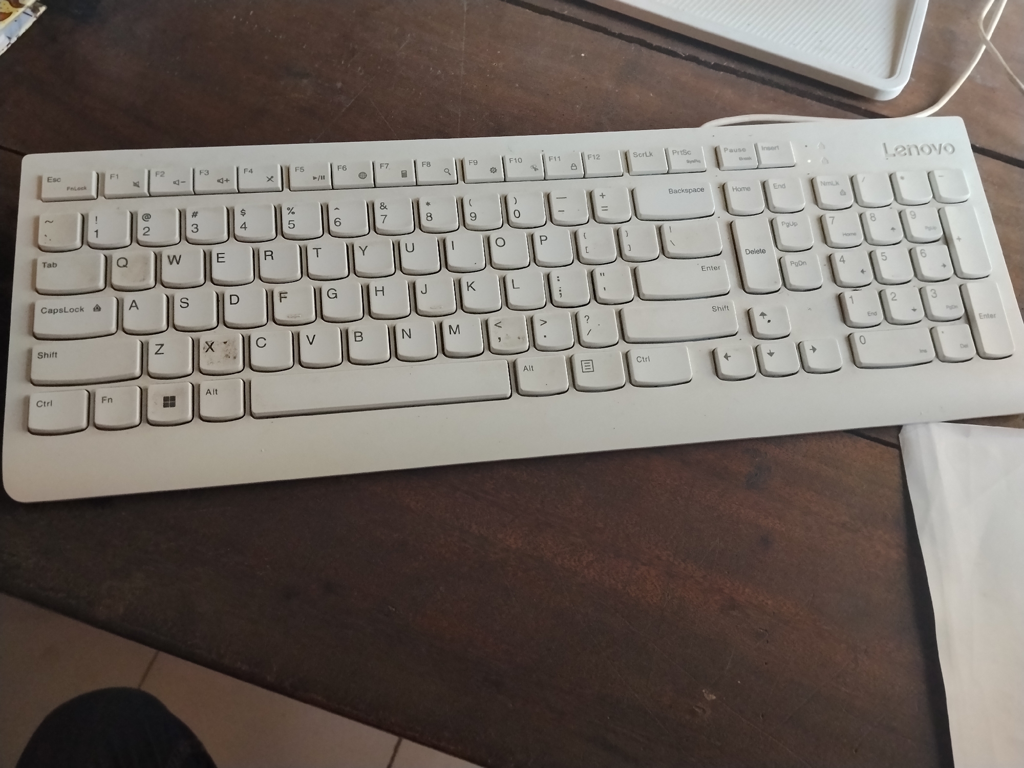 A white Lenovo keyboard is placed on a dark wooden table. The keyboard is full-sized and includes a numeric keypad. The keys are clean and show no signs of wear. The image is taken from a slightly elevated angle, looking down at the keyboard. There is no specific information about the city of Laok Jangjang, Indonesia, or any people present in the image. The environment appears to be an indoor setting, likely a home or office, given the presence of the keyboard.