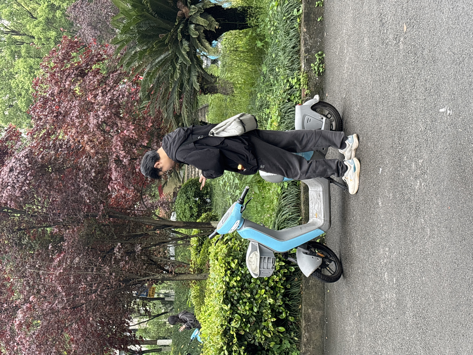 A person is standing beside a light blue scooter on a paved path. The person is dressed in black clothing and is looking down at something in their hands. The scooter is parked on the side of the path, with greenery and trees in the background. The location is Jiangjin, China.