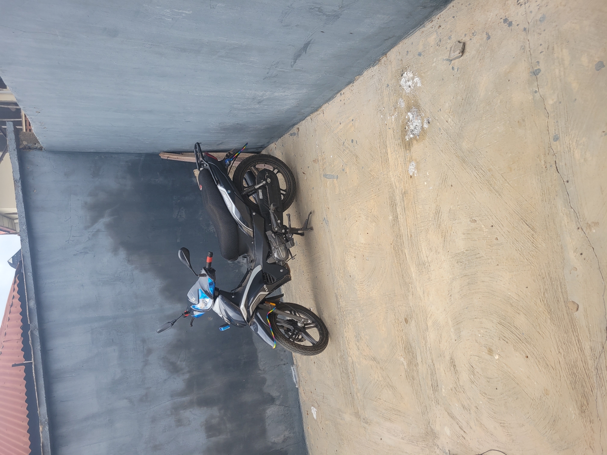A black and white motorcycle is parked on a concrete surface in an outdoor courtyard. The motorcycle is angled towards the viewer's right, with its front wheel turned slightly to the left. It has a black seat, silver accents, and blue and white detailing on the front fairing. The courtyard is enclosed by gray concrete walls. A red-tiled roof is visible in the background at the top of the image. The concrete ground is rough and textured, with some white debris scattered in places. A wooden plank leans against the wall behind the motorcycle. The scene appears to be in an urban or suburban environment.