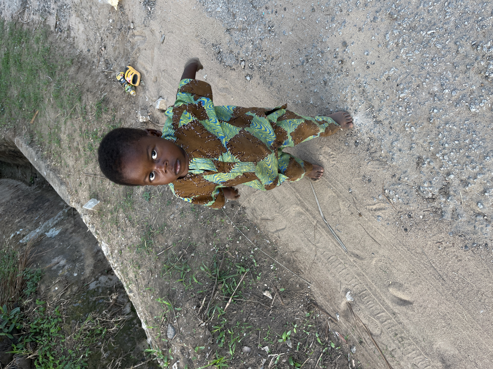 A young child stands barefoot on a dirt and gravel road in what appears to be a partially developed or rural area. The child wears patterned brown and green clothing and looks up toward the camera. A toy vehicle sits on the ground near the child's left. To the left of the child is a concrete structure and vegetation. Tire tracks are visible on the dirt and gravel road. The image appears to be taken outdoors in daylight. It is not possible to confirm the location is Abuja, Nigeria from the image alone.