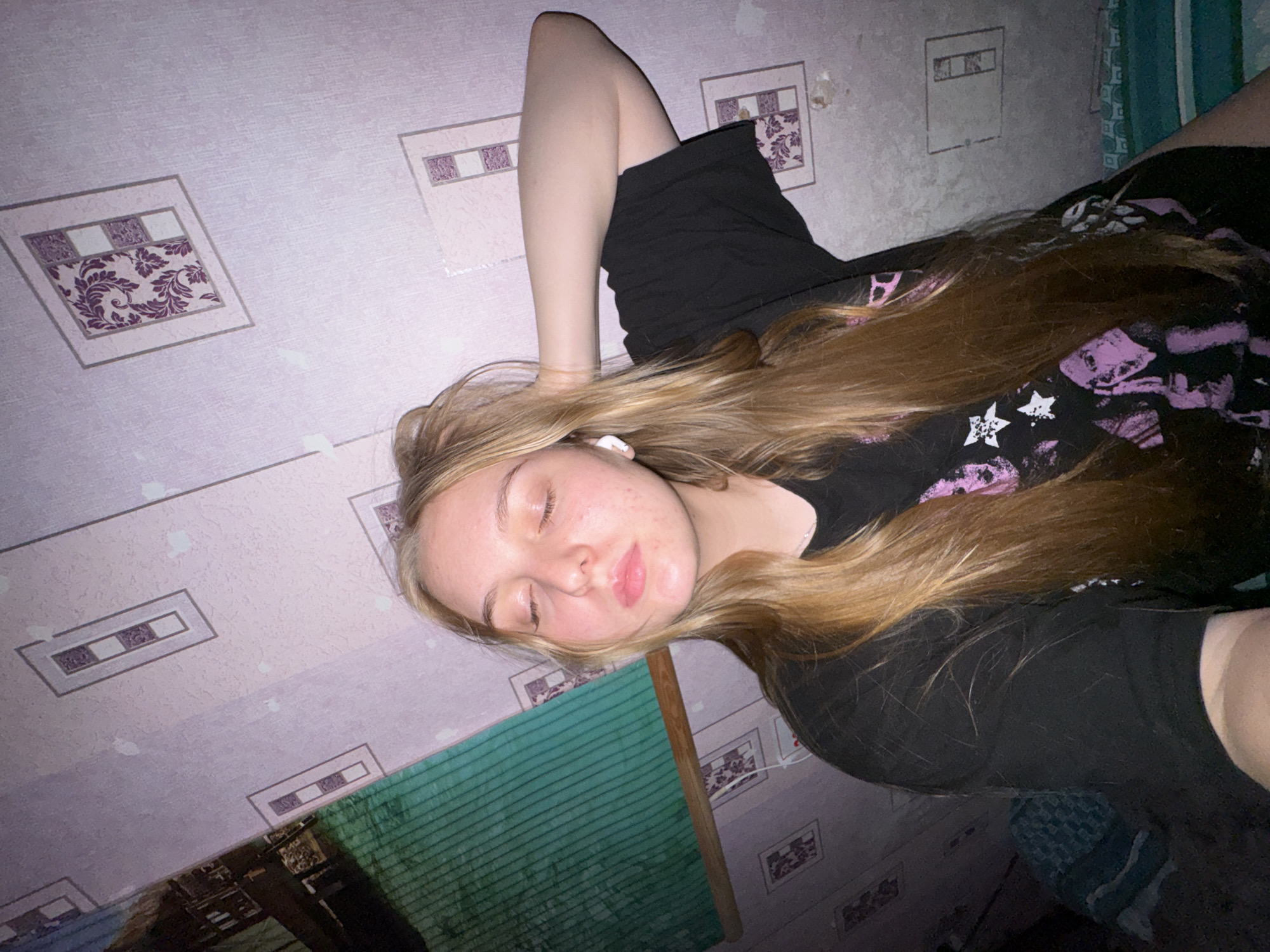 In the provided image, a young woman with long blonde hair is depicted with her eyes closed, one arm behind her head. She is wearing a black t-shirt with a graphic print. She has earbuds in her ears. The background consists of a wall with a pinkish-purple wallpaper featuring a repeating pattern of rectangular frames, some containing floral motifs and others a textured pattern. To the left of the woman, a portion of a green, textured surface with wooden trim is visible, which appears to be a window blind or screen. The lighting suggests an indoor setting, possibly at night or in a dimly lit room. The overall impression is of a casual, personal moment captured in a private space. This image does not contain specific visual cues to pinpoint a location within the city of Perm, Russia.