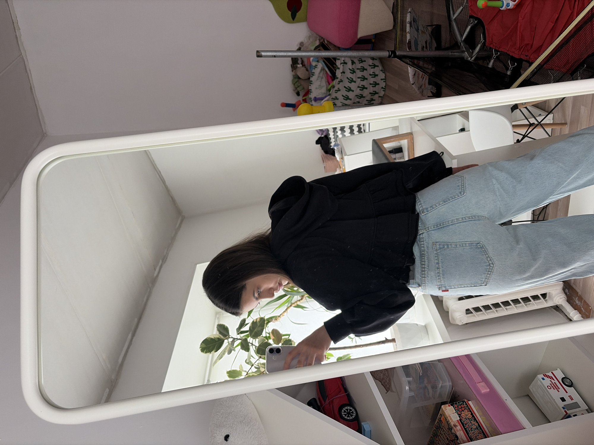 A person wearing a black hoodie and light blue jeans is taking a mirror selfie. They are holding a smartphone. The reflection in the mirror shows a room interior with a plant, a white shelving unit with various items, and part of a wall. Other objects, including a child's trampoline, are also visible in the background. There is no verifiable information indicating this photo was taken in Chaikovsky, Russia.