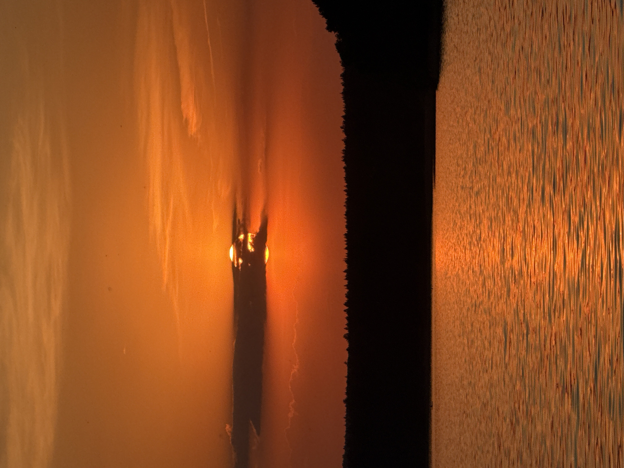 The image depicts a sunset over water. The sun, visible as a bright orb, is partially obscured by clouds or a silhouetted landmass. The light from the sun is reflected on the surface of the water, creating a golden shimmer. The sky has a gradient of orange and yellow hues, suggesting the time of day. A dark, undefined landmass is present on the right of the image. The exact location within Votkinsk, Russia, cannot be determined with certainty from the image alone, but it is likely taken from a vantage point with a view of a body of water, possibly the Votkinsk Reservoir or a similar water feature within the city. No people are visible.