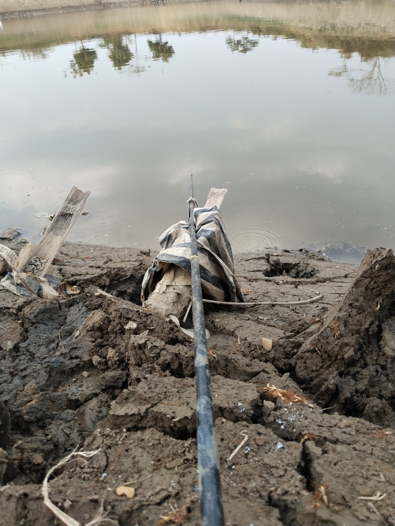 The image depicts a close-up, low-angle shot of a muddy bank beside a body of water. A fishing rod extends into the frame from the bottom right, angled towards the water. The bank is composed of cracked, dark brown mud and debris, including a piece of weathered wood leaning precariously. Wrapped around a central object on the bank is a piece of striped fabric, possibly a tarp or bag. The water reflects trees and the sky, creating a soft, hazy effect. The overall scene suggests an outdoor, possibly abandoned or neglected, fishing spot.