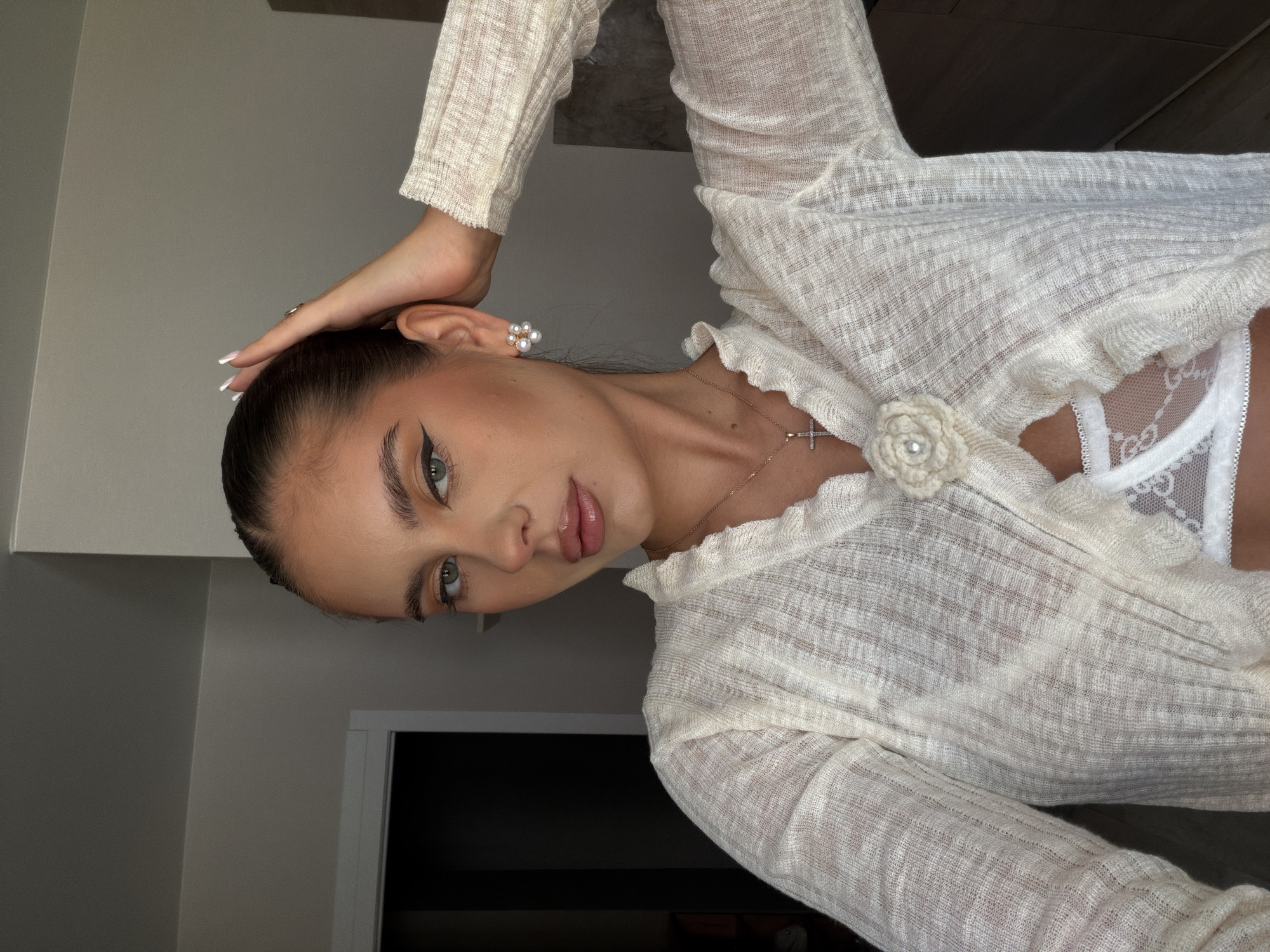 A young woman poses indoors, wearing a light, knitted, long-sleeved cardigan with a flower-shaped pin. A glimpse of a white, lacy garment is visible underneath. She has her hair pulled back and is wearing pearl earrings and a delicate necklace with a cross pendant. She is looking directly at the camera, with one hand resting on her head. The background is a neutral-colored wall with a doorway visible in the lower-left corner. There is no explicit evidence to place this image in Krasnoyarsk, Russia.