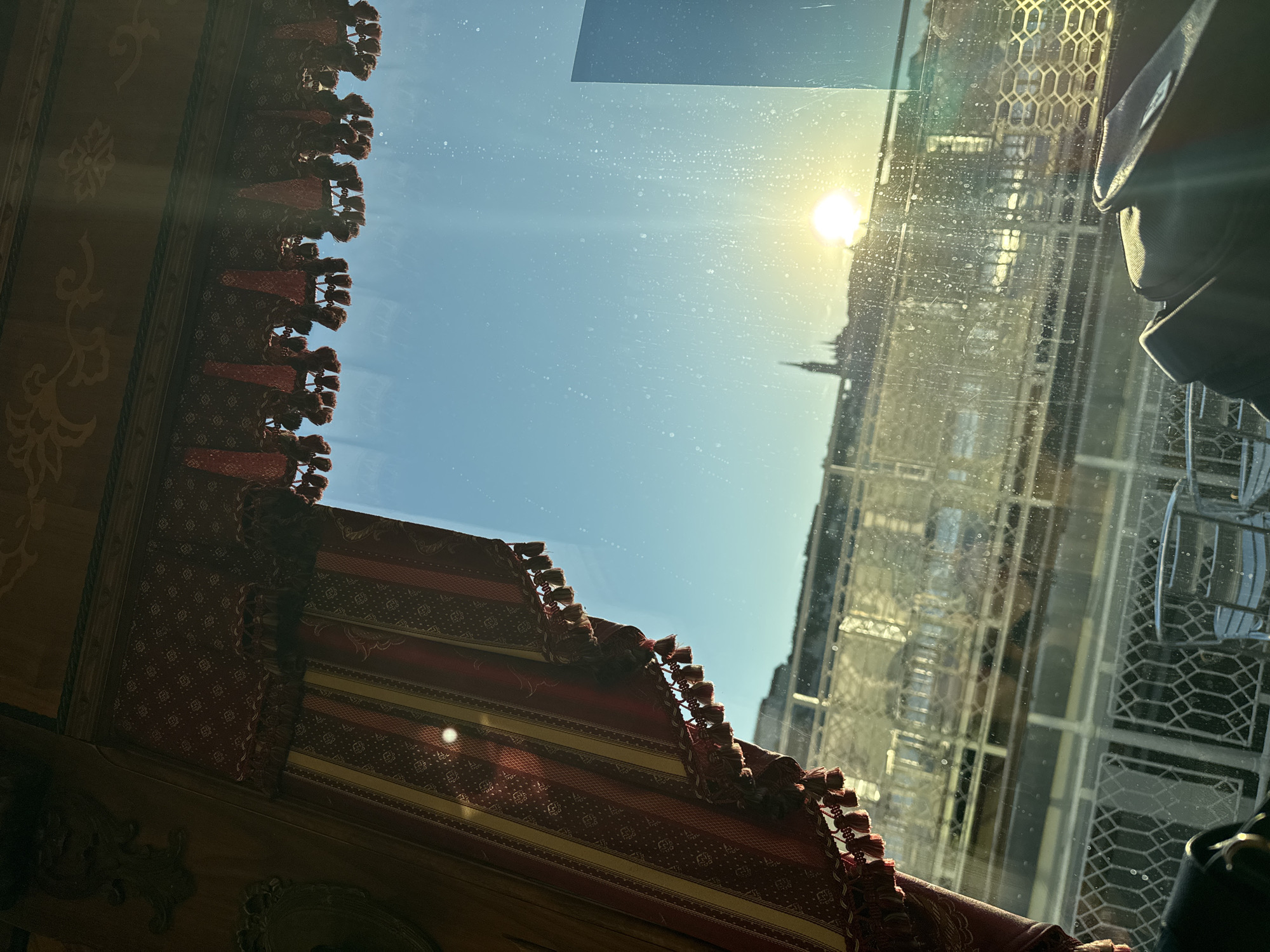 The image captures a bright, sunlit view taken from inside a structure, looking outwards through a window.
**Subjects:**
In the foreground, on the left and extending along the top and bottom edges, is elaborate interior decoration. This includes richly patterned red and gold fabric adorned with tassels, possibly part of a curtain, valance, or upholstery. Dark, ornate wooden panels with intricate scrollwork are also visible on the far left.
Through the window, the central and right portions of the image are dominated by a clear, vibrant blue sky. A powerful, bright sunburst is prominent on the upper right, casting a strong flare. The windowpane itself appears to have numerous small dust or water specks scattered across its surface.
Reflected prominently on the right side of the glass, a tall building with a distinctive, complex geometric lattice pattern on its facade is visible. Further in the distance, partially silhouetted against the sun, is a faint outline of a pointed structure, possibly a minaret or a spire. A dark, padded object, resembling a bag or part of an interior seat, can be seen in the lower right corner, possibly another reflection or part of the immediate interior.
**Activity or event taking place:**
The image presents a static scene, capturing a momentary view from an indoor space looking out. There is no specific activity or event taking place, but rather an observation of the exterior world from a well-decorated interior.
**Notable details:**
The time of day appears to be daytime, with a clear sky and intense sunshine. The strong glare from the sun suggests direct sunlight. The intricate interior decor, combined with the distinctive geometric architecture reflected in the window (possibly including a minaret), hints at a location with rich cultural or Islamic architectural influences. No emotions are explicitly conveyed.
**Visible text:**
There is no visible text in the image.