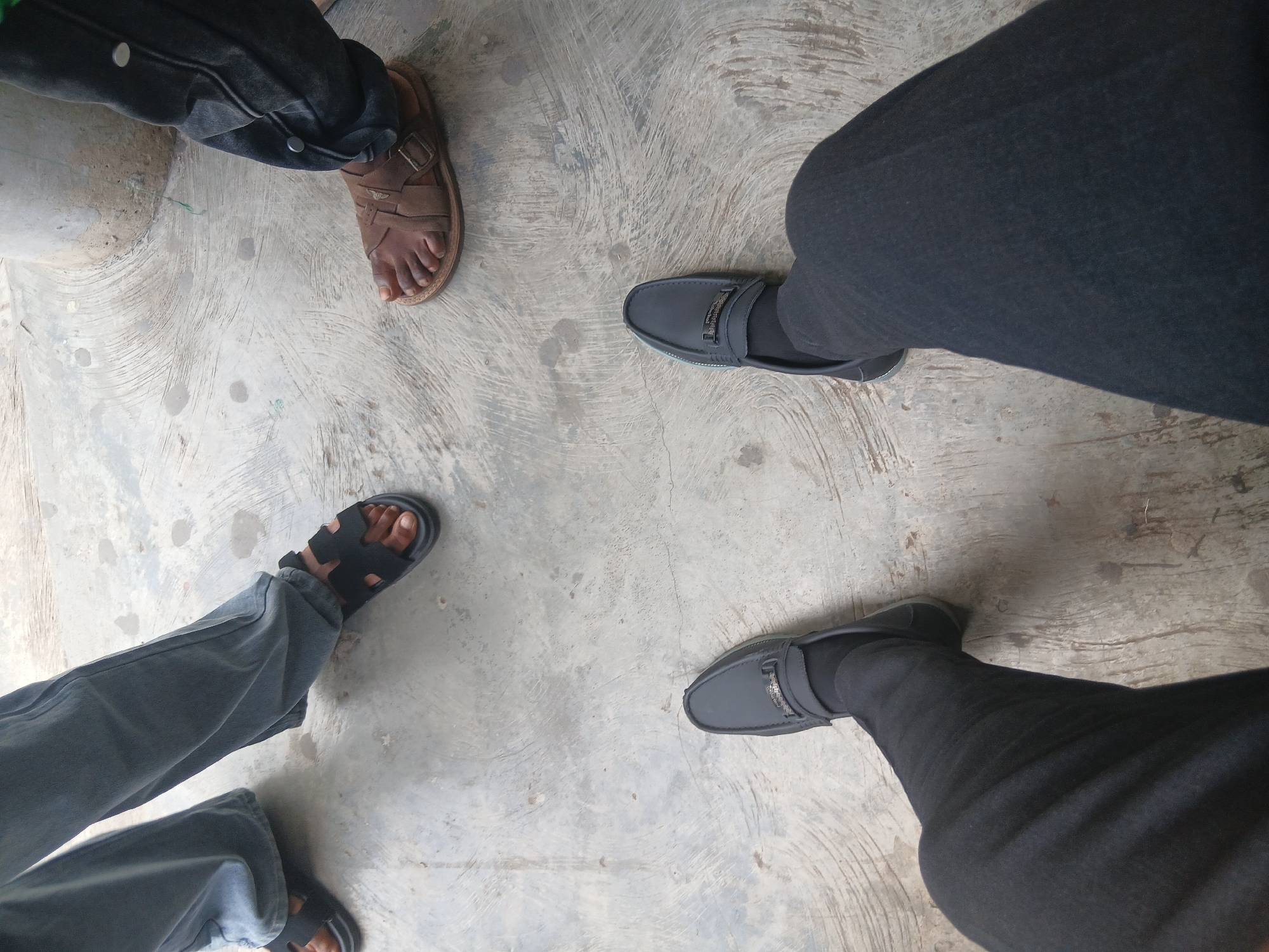 The image shows four pairs of legs and feet standing on a concrete surface with a circular pattern. The legs are arranged in a rough circle. The top-left leg is wearing dark-colored jeans and a brown sandal. The top-right leg is wearing dark-colored pants and black leather shoes with visible socks. The bottom-left leg is wearing blue jeans and black sandals. The bottom-right leg is wearing black pants and black leather shoes with visible socks. The scene appears to be indoors or on a covered surface. The location is presumed to be Lagos, Nigeria.