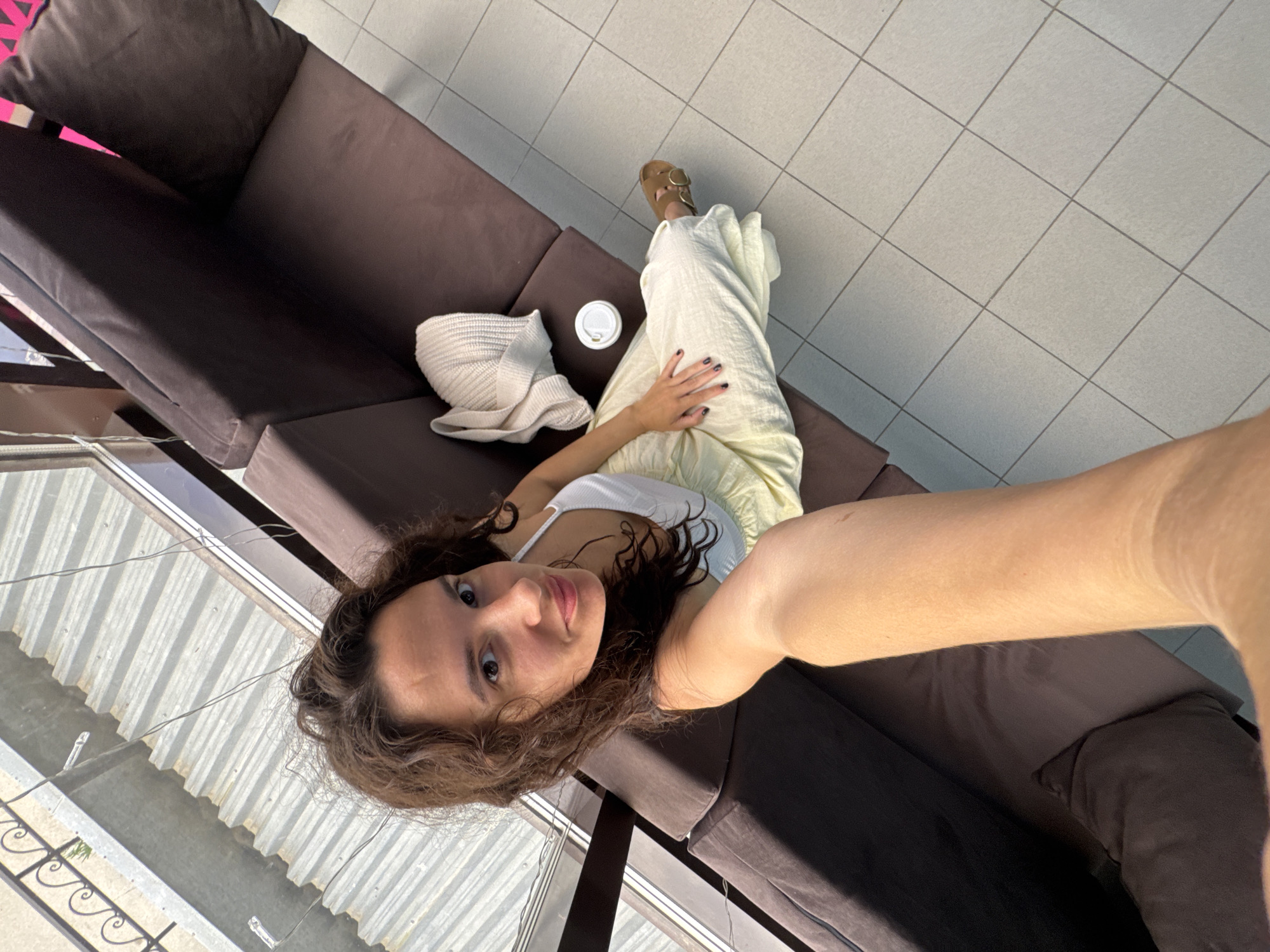 A young woman is taking a selfie while lying on a dark brown couch. She is wearing a white tank top and loose, light yellow pants. Her hair is dark brown and curly, and she is looking up at the camera with a relaxed expression. To her left, on the couch, are a white knitted bag and a white paper cup with a lid. The couch is situated in an indoor setting with light-colored tiled flooring. Sunlight streams in from the left side of the image, casting shadows on the couch and illuminating the woman's arm and face. The overall mood is casual and serene.