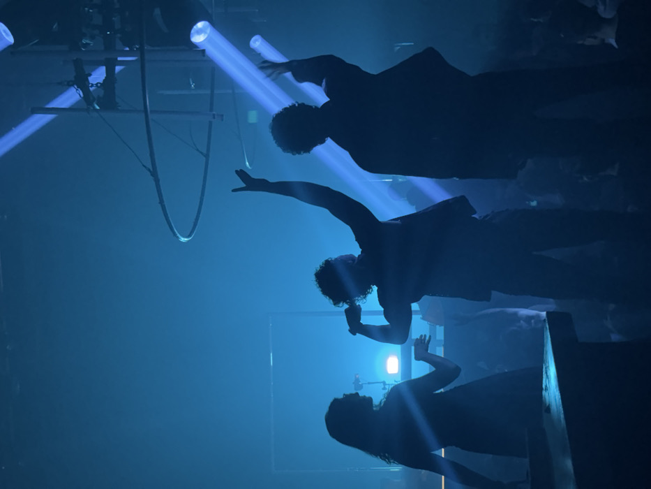 Silhouetted figures stand under blue stage lighting. Two men are visible above, one with an arm raised. Below them, a woman raises her hands slightly. A square frame stands behind them, illuminated by a bright light. Above, cylindrical lights shine blue beams down onto the scene. Hanging from the ceiling are rigging mechanisms. The overall mood is dramatic and theatrical, likely depicting a performance.