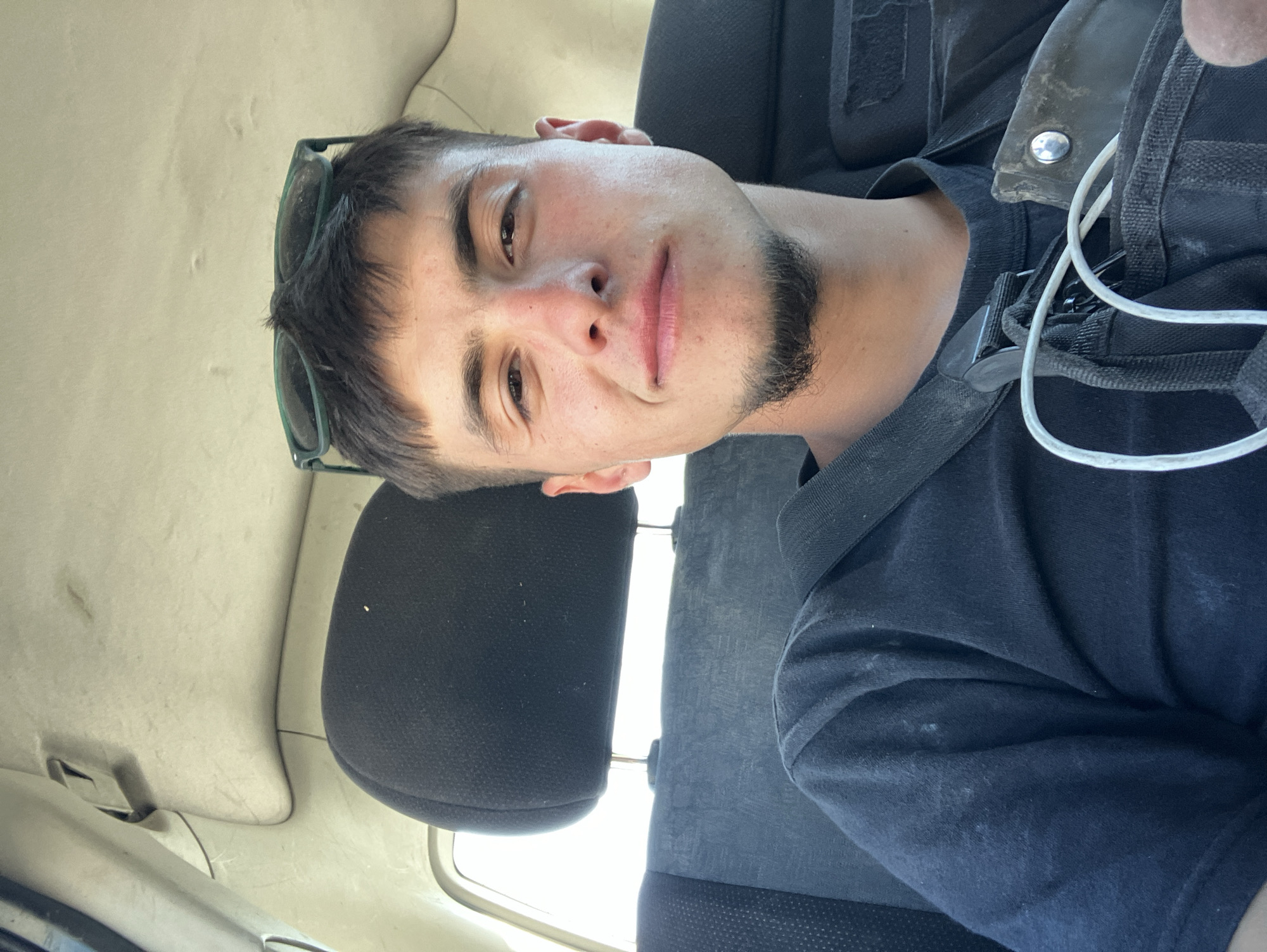 A young man is seated in a car. He is facing the camera, and sunglasses rest on his head. The man has a short, dark beard. He wears a dark t-shirt and a dark vest with white cords hanging from it. The interior of the car is light-colored except for the dark seats. The location is unspecified but assumed to be Novo Selo, Bulgaria.