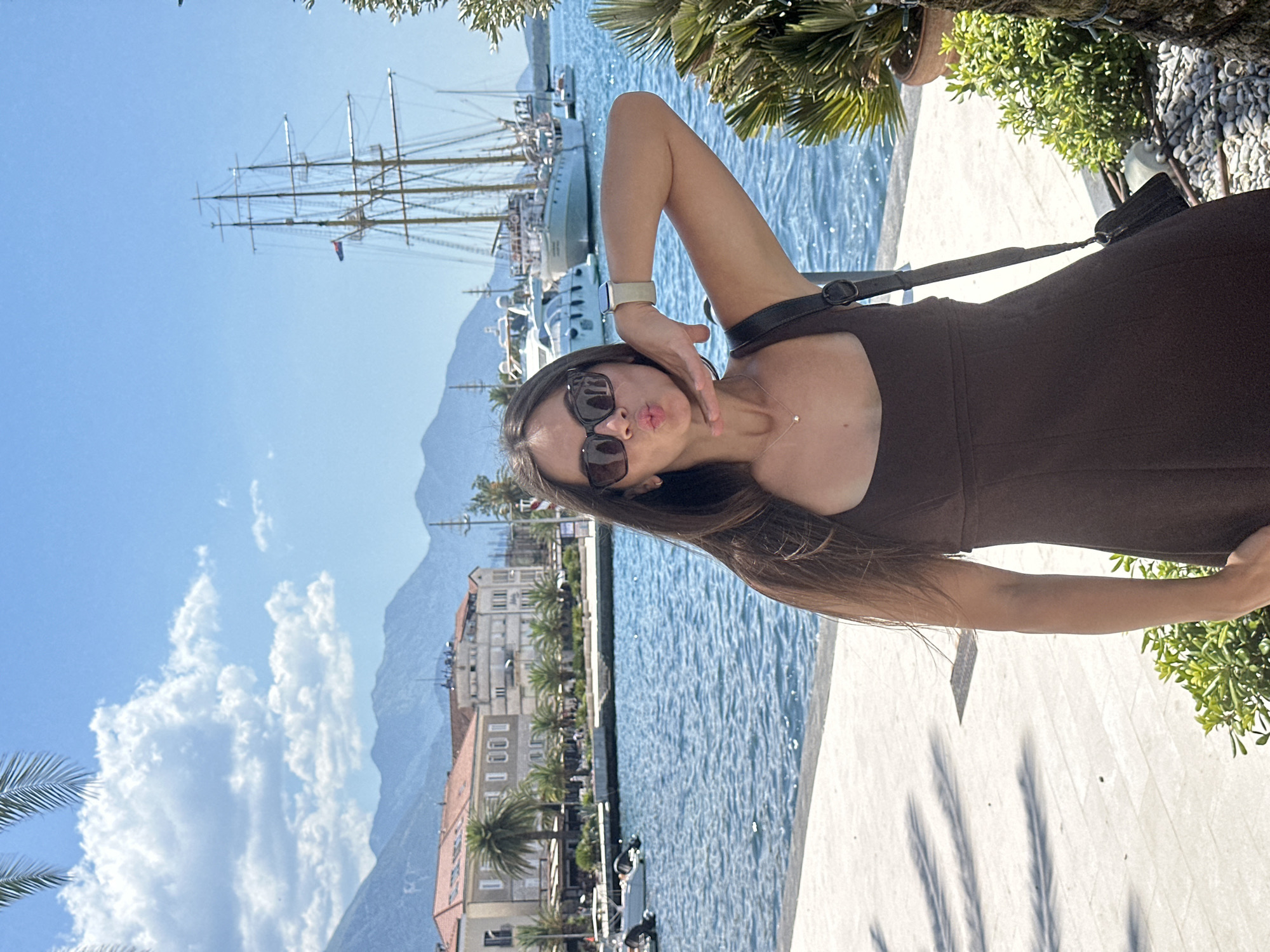 A young woman with long, dark hair and sunglasses poses for a selfie on a paved walkway beside a body of water. She is wearing a dark brown dress and a small dark purse is slung over her shoulder. Her right arm is raised with her hand near her face, and she is puckering her lips. Behind her, the water is blue and choppy, with several boats docked, including a large sailing ship with multiple masts. In the background, buildings with red tile roofs and distant mountains are visible under a bright blue sky with scattered white clouds. The scene appears to be in a coastal city, likely Donja Lastva, Montenegro, given the architectural style and the presence of palm trees, which can be seen in the foreground and middle ground.