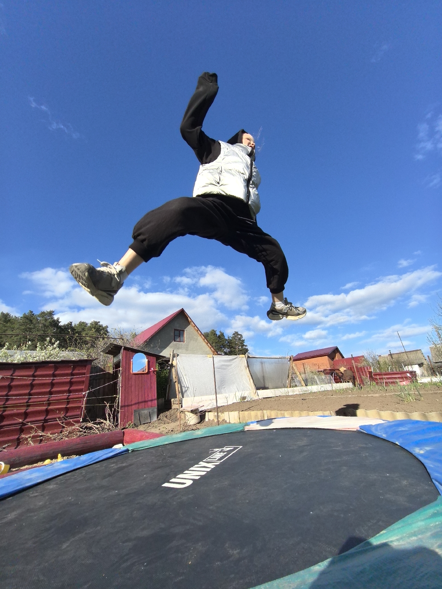 A person is in mid-air above a trampoline. The person wears dark-colored pants, a light-colored vest, and sneakers. One arm is raised. In the background are rural-style buildings, including a house with a red roof and a red-walled structure. A garden area with rows is also visible. The sky is blue with scattered clouds. The trampoline is labeled "UNIX." The location is Noviy, Russia.