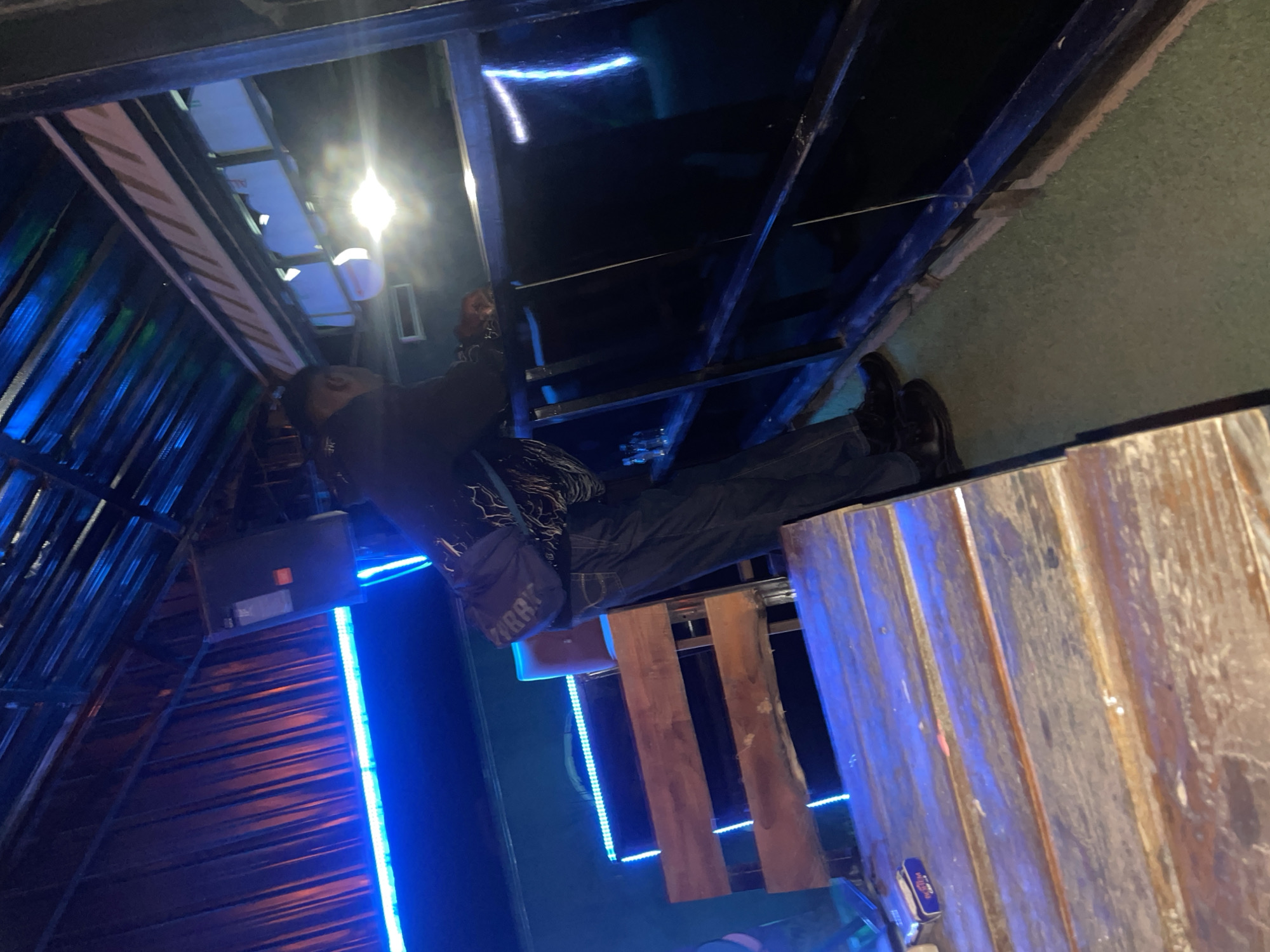 A man in dark clothing stands inside a structure with blue LED lighting. The structure appears to be under construction or partially assembled, with visible metal framing and wooden planks. The environment is dimly lit, and the perspective is tilted, suggesting a photograph taken from an unusual angle. The man's face is obscured by shadow. Gura Top, Nigeria, is not discernible from the image alone. The image contains a person, building structure, and LED lights.