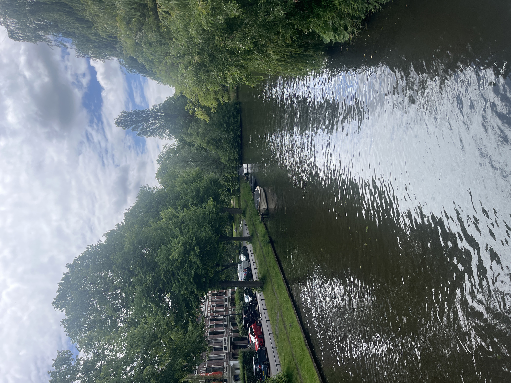 A high-angle medium shot captures a canal in a city, bordered by a tree-lined street with parked cars and buildings on one side, and lush vegetation on the other. The canal's dark water reflects the overcast sky and surrounding greenery, creating a textured surface. Along the left bank, a row of red-brick buildings with tall windows faces a grassy verge where several cars, including a red hatchback, are parked. A person on a bicycle rides past the parked cars. Further along the canal, two small boats are moored. The right bank is densely covered with trees, including weeping willows. The sky is a mixture of grey clouds and patches of blue.