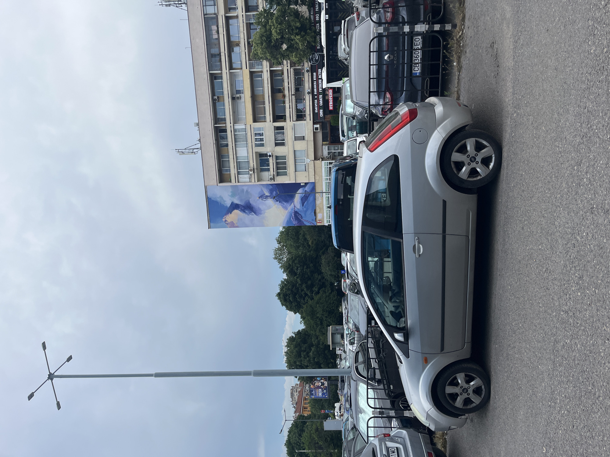 The image depicts an outdoor scene in a city, likely Sofia, Bulgaria, with a focus on a parking lot and a multi-story building. In the foreground, a silver three-door hatchback car, identified as a Ford Fiesta based on its design, is parked. Behind it, other vehicles are visible, including a dark-colored Renault Megane with a license plate reading "CB 360 EO". The parking lot is paved with asphalt.
The background is dominated by a light-colored, multi-story apartment building with numerous windows and balconies. A large mural, featuring a depiction of a figure in a snowy, mountainous landscape, is painted on the side of the building. Several businesses appear to be located on the ground floor of the building, with signage indicating food and street food services.
A tall street light with four lamps stands to the left of the frame. Trees line the area behind the parking lot and in front of the building, suggesting a green urban environment. The sky is overcast with a muted, cloudy texture. The overall impression is of a typical urban setting with residential and commercial elements.