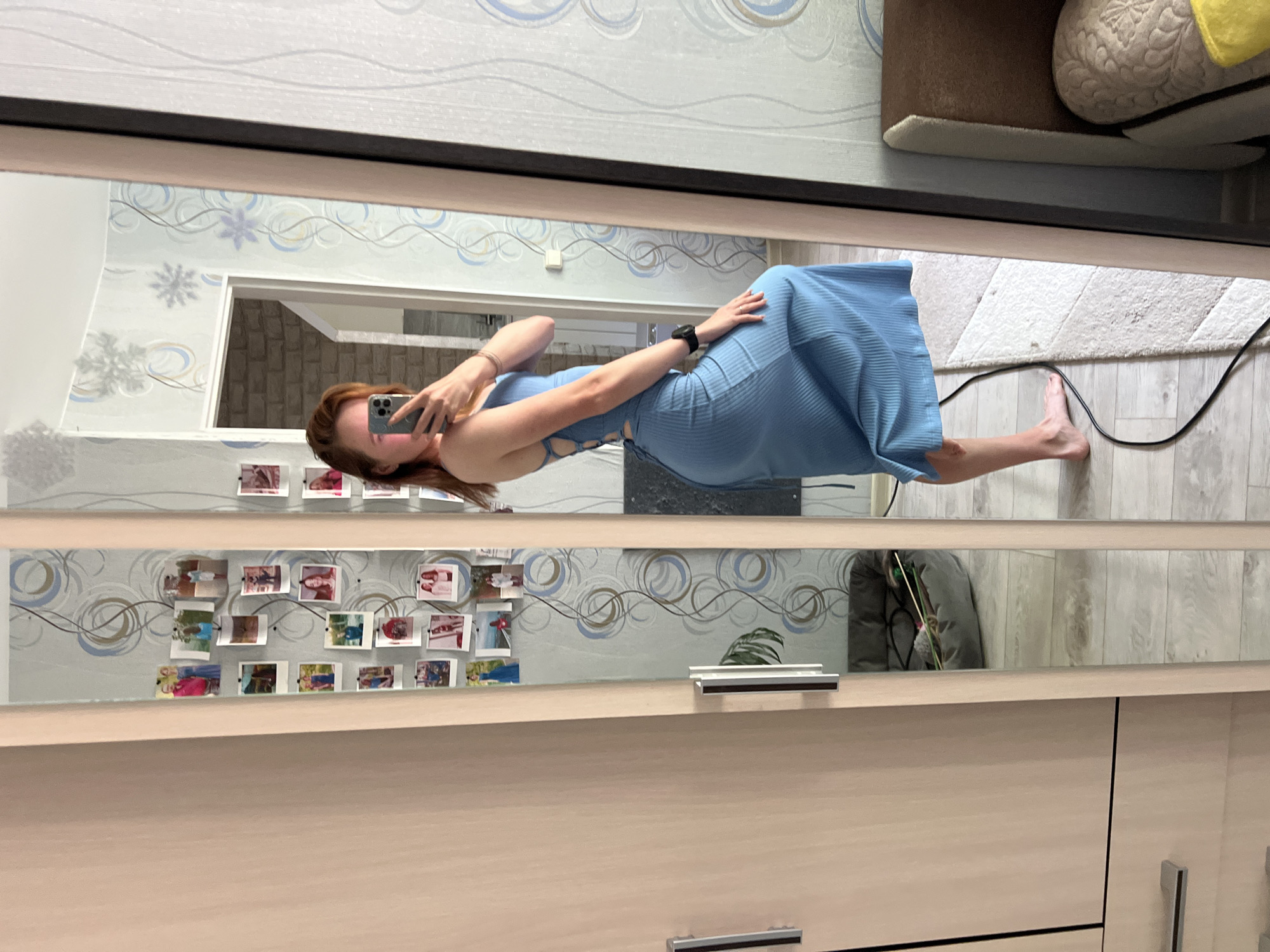 A woman is taking a selfie in front of a mirror in what appears to be an apartment. The woman has red hair and is wearing a blue dress. She is holding a phone in her left hand and is looking at the camera. The mirror is reflecting the room behind her, which includes a doorway, a wall with photos, and a piece of furniture. The location is Chaikovsky, Russia.