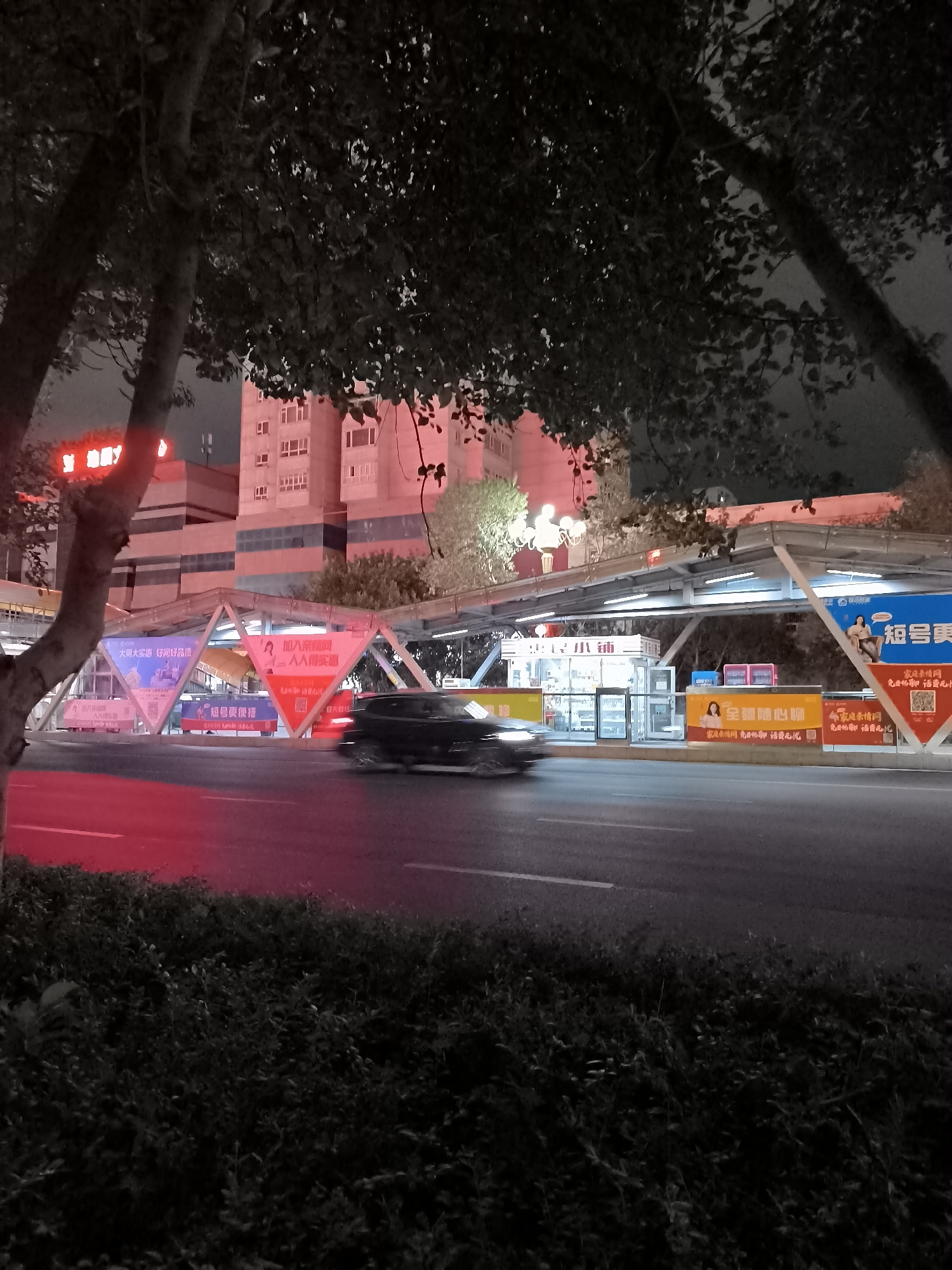The image depicts a street scene at night in Ürümqi, China. A pedestrian overpass with advertising banners is visible spanning a road. A black car is in motion on the road, blurred due to movement. The overpass is illuminated, and the surrounding buildings are lit with red and white lights. Trees with dark foliage are present in the foreground. The scene appears to be an urban environment with commercial activity indicated by the signs and illuminated structures.
