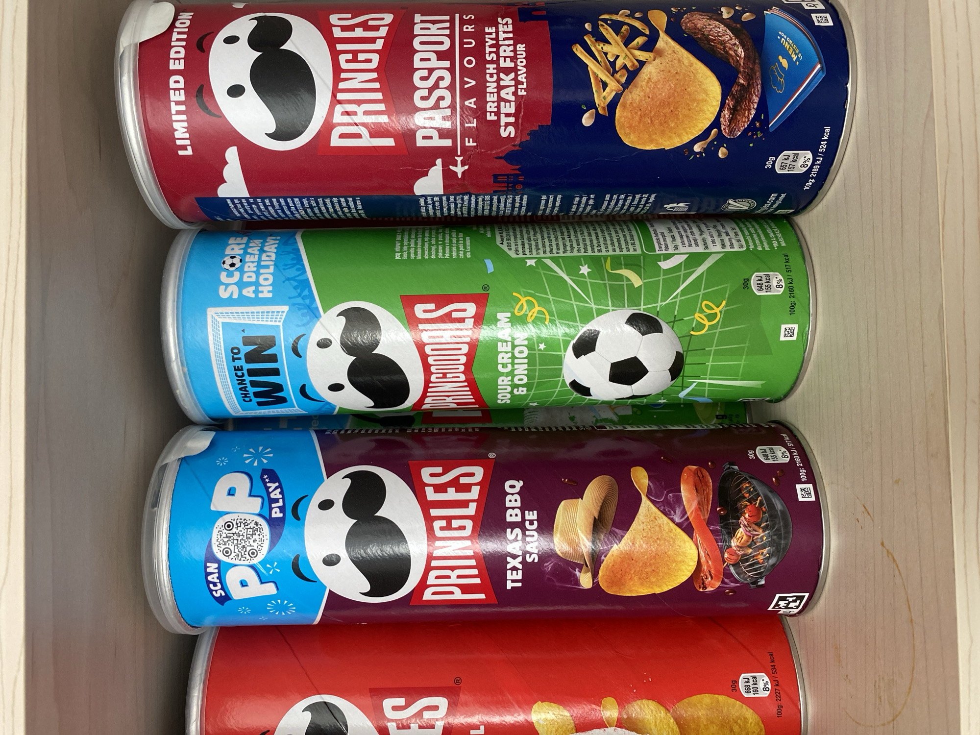 A close-up, eye-level, interior shot shows four cans of Pringles potato chips stacked side-by-side on a light-colored wooden shelf. The cans are arranged from left to right, with the leftmost can partially visible and red in color, featuring white text. The second can from the left is blue and purple with a "Scan POP PLAY" graphic and an image of a Pringles mascot. The third can is green and blue, with "PringoGOALS" and "Sour Cream & Onion" text, also featuring a soccer ball graphic and a Pringles mascot. The fourth can, on the far right, is red and blue and labeled "LIMITED EDITION Pringles PASSPORT FLAVOURS FRENCH STYLE STEAK FRITES FLAVOUR," also with a Pringles mascot. The cans are in focus, and the wooden shelf shows some faint brown circular stains. There are no people or animals present in the image. The scene appears to be a retail shelf or a home pantry. There is no information within the image to indicate its location in Chișinău, Moldova.